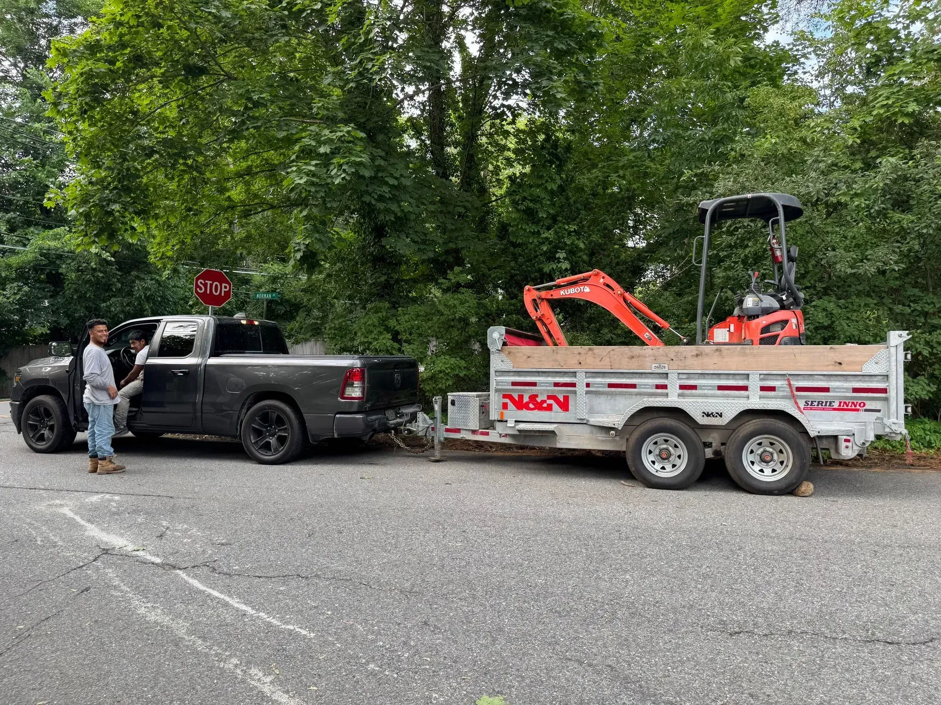 A black truck pulling a trailer with a small excavator parked on a paved road. A person stands at the truck's open door.