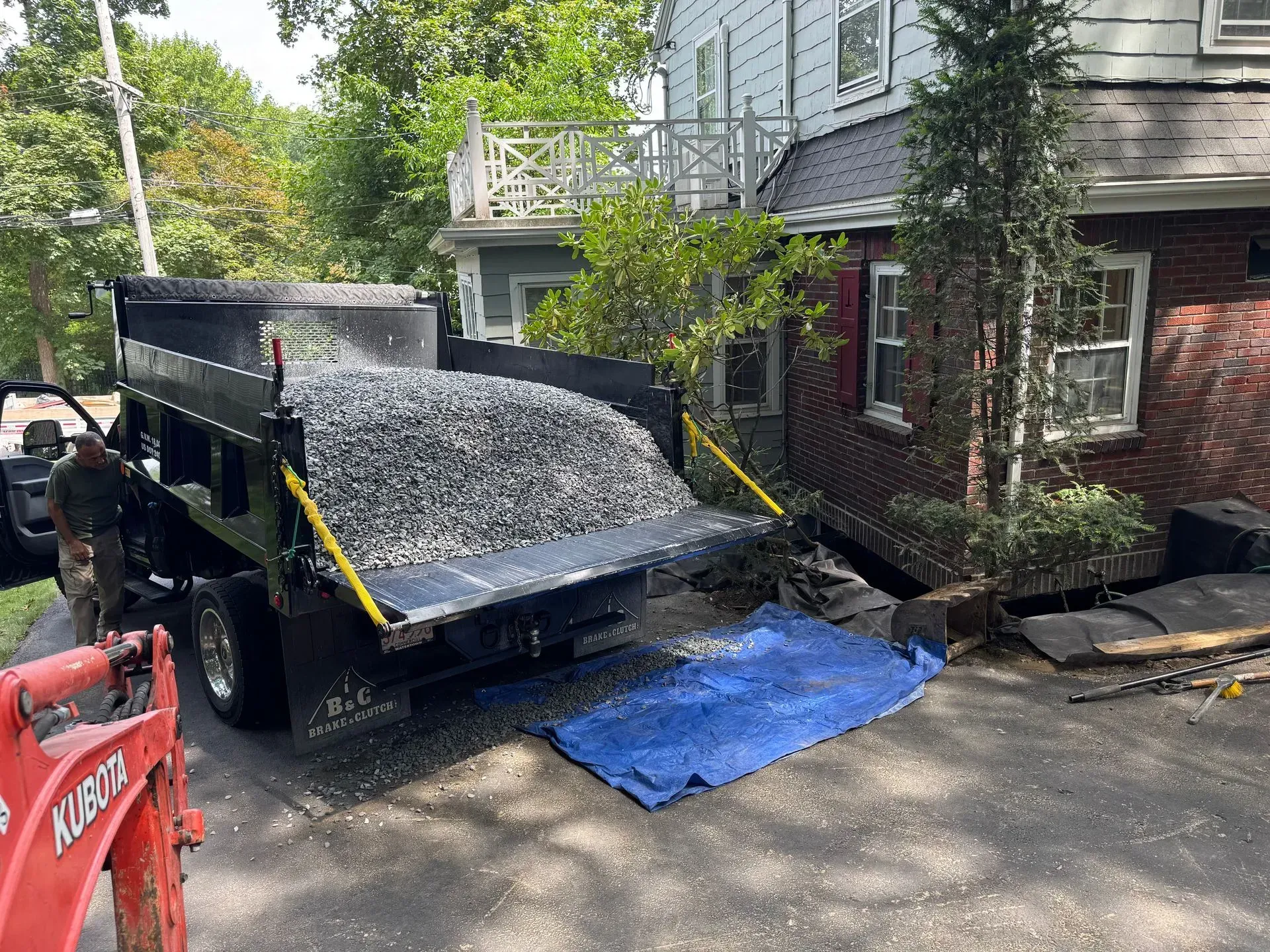 Dump truck unloading gravel near a house. A worker stands nearby.