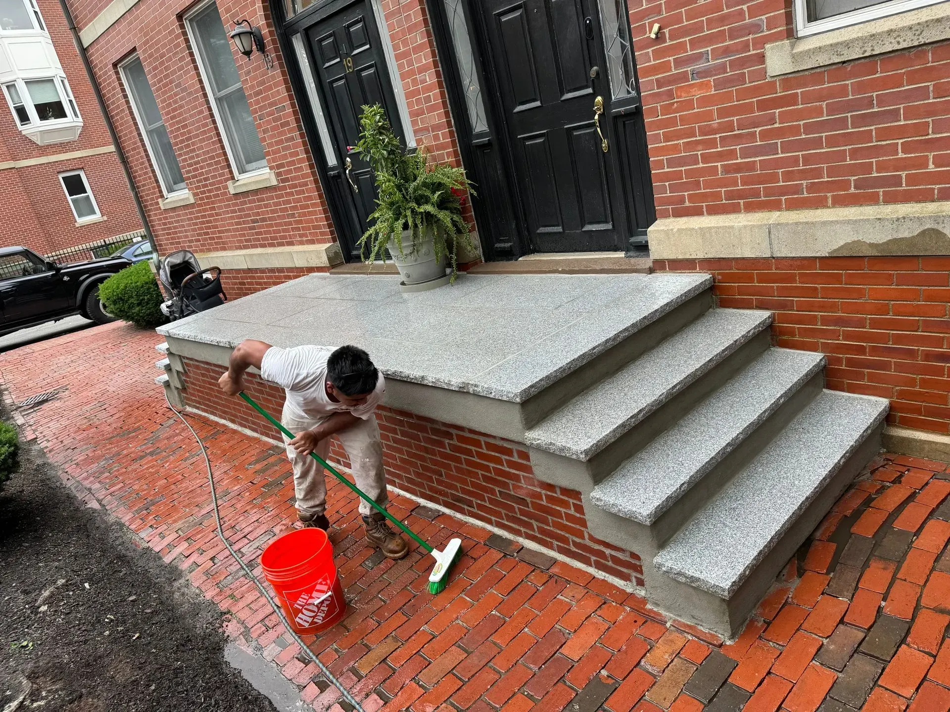 Man sweeping steps outside a brick building with a green-handled brush and red bucket. Grey steps and entrance.