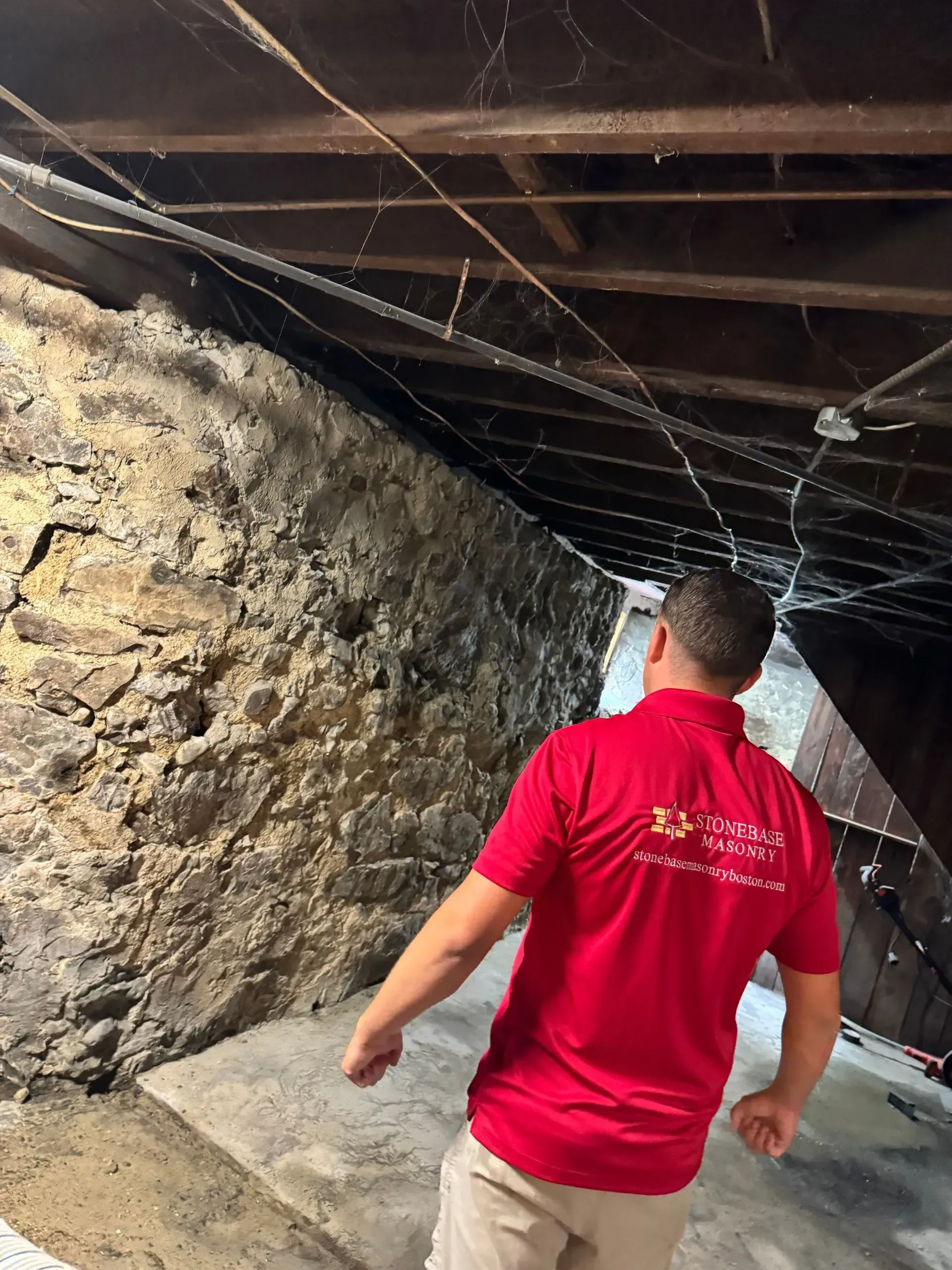 Man in red shirt inspecting a stone foundation basement. Spiderwebs, exposed wiring, and wooden beams are visible.