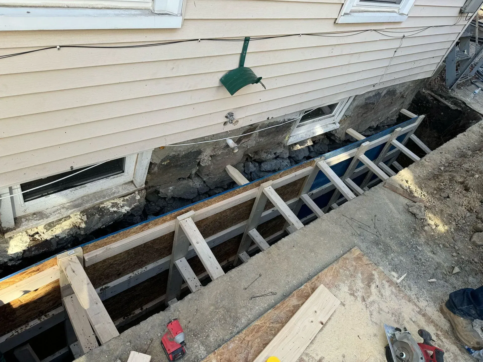 Construction site; excavated area under a house with wood framing and a broken wall section, debris present.