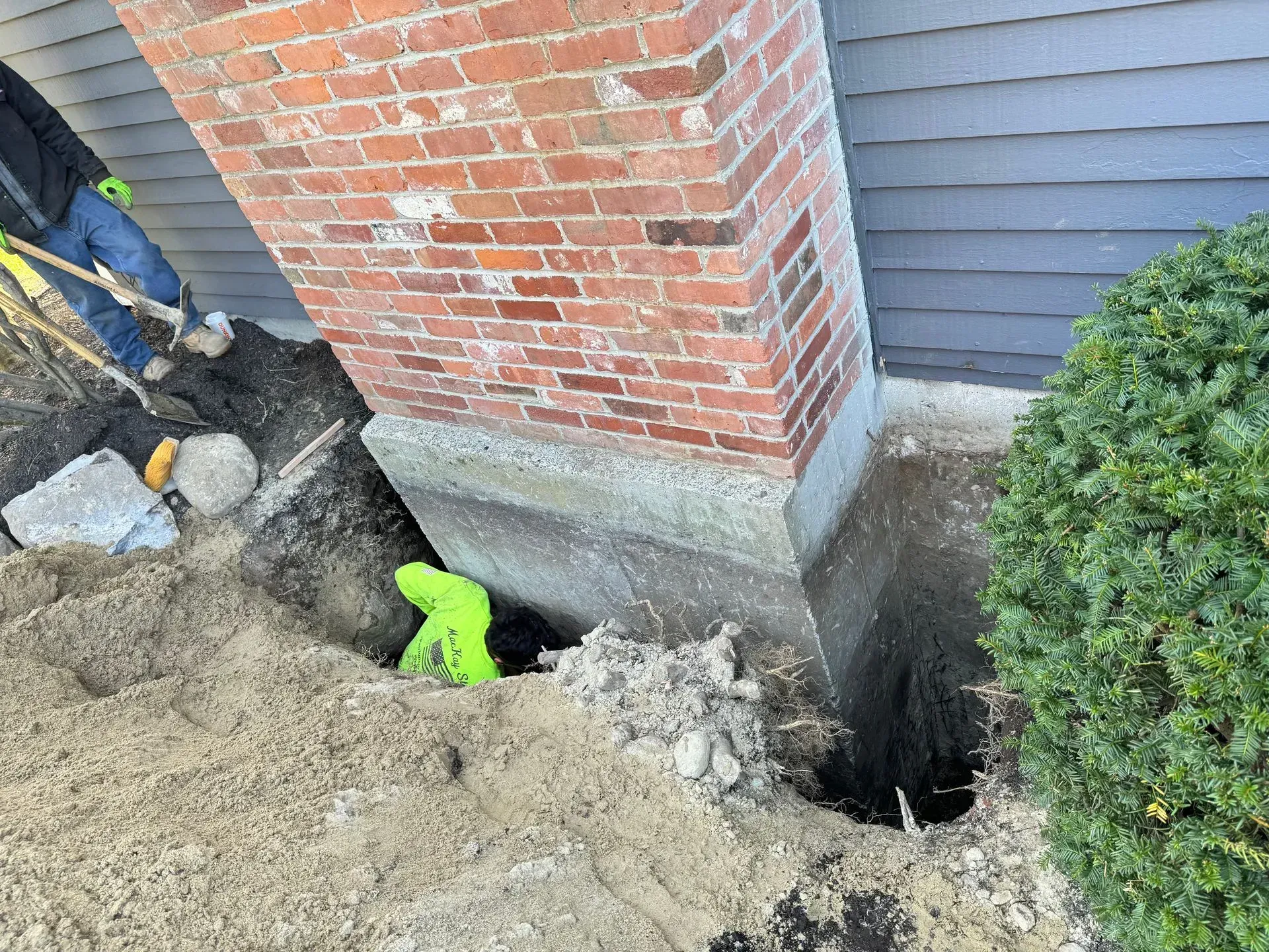 Excavation around a brick chimney with concrete base. A person is working in the trench wearing green gloves.