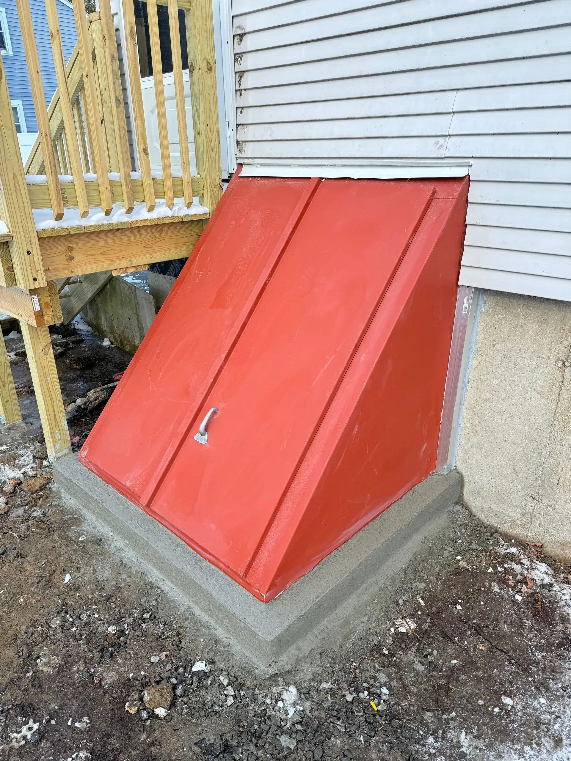 Red basement bulkhead door set in a concrete foundation next to wooden stairs and a white house.
