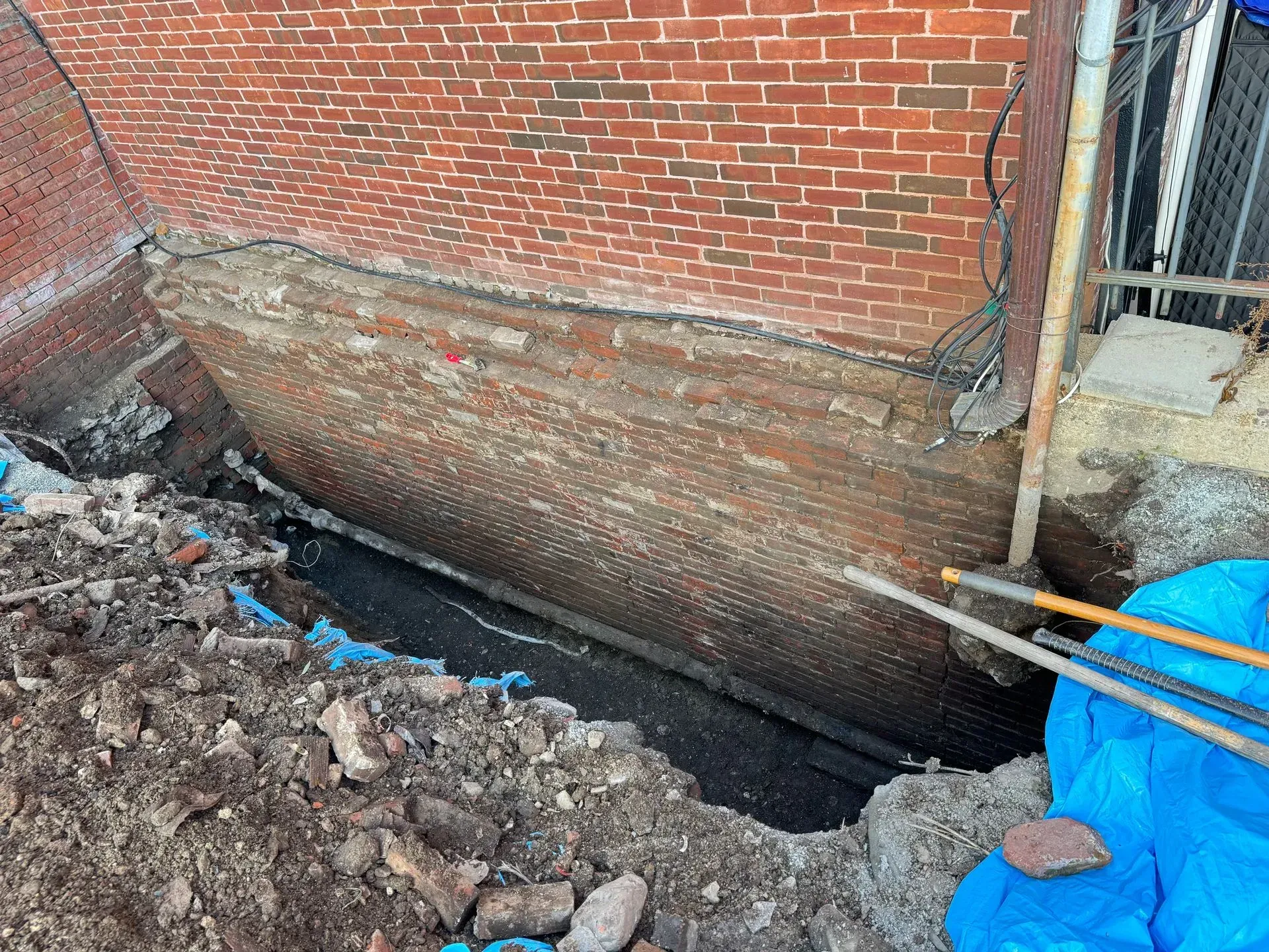 Excavated brick foundation of a building; dirt and debris surround the base; blue tarp visible.