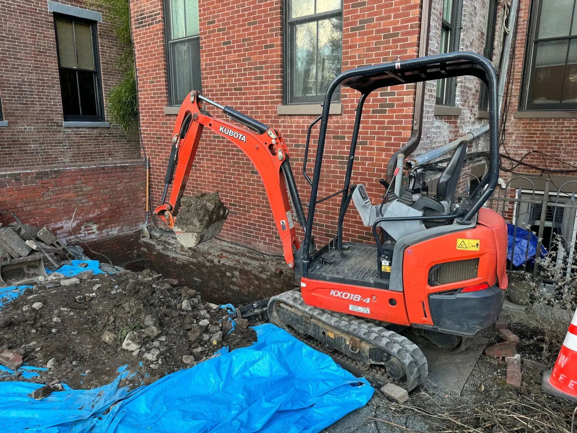 Orange Kubota excavator digging next to a brick building. Blue tarp and debris visible.