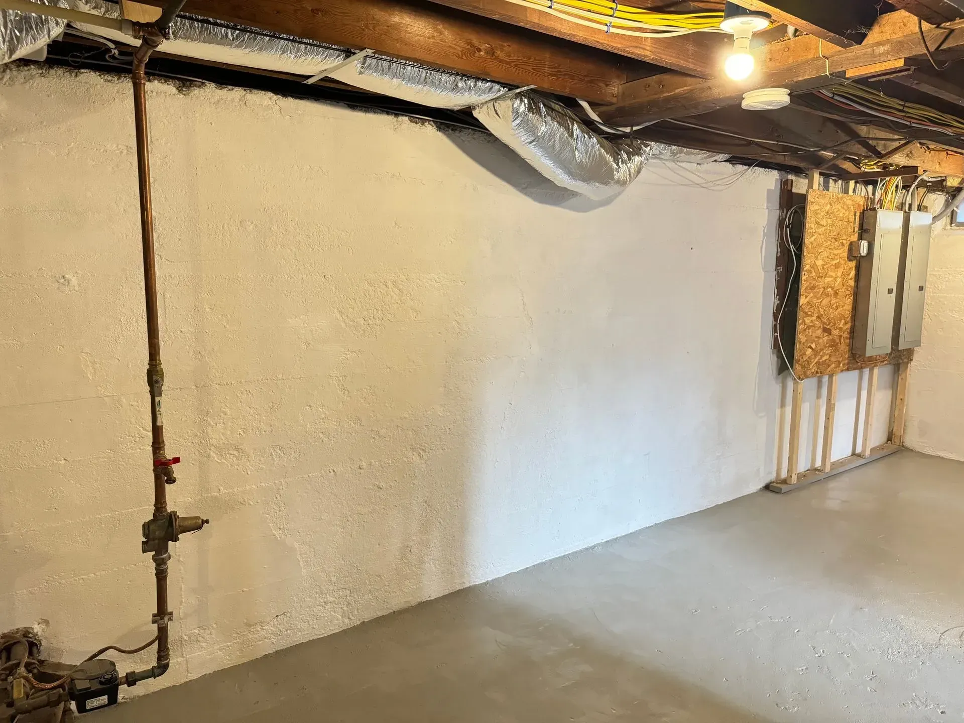 Basement wall with spray foam insulation, exposed beams, and concrete floor. Plumbing and electrical box visible.