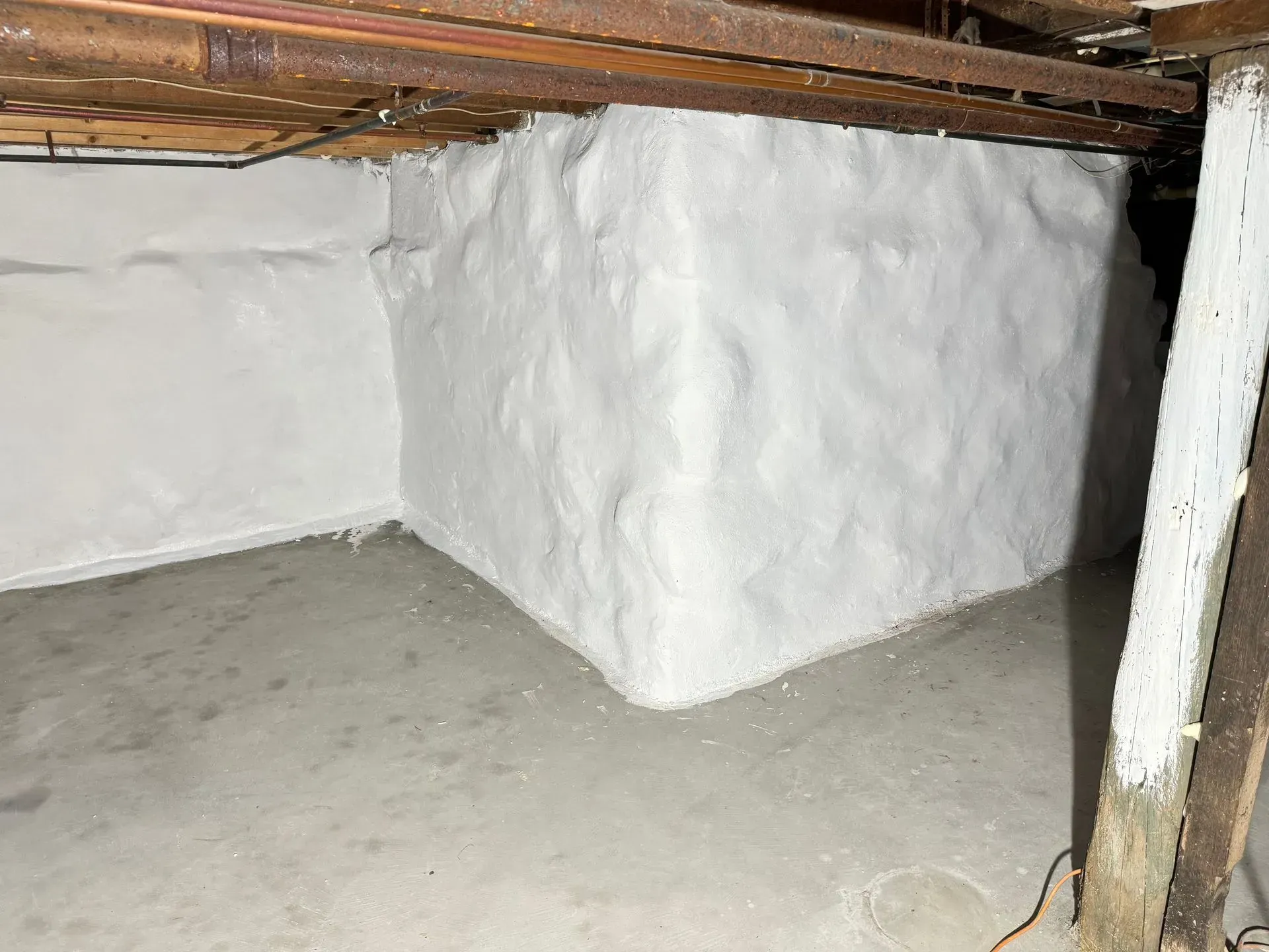 White-painted foundation corner in a basement. Concrete floor with exposed wooden beams overhead.
