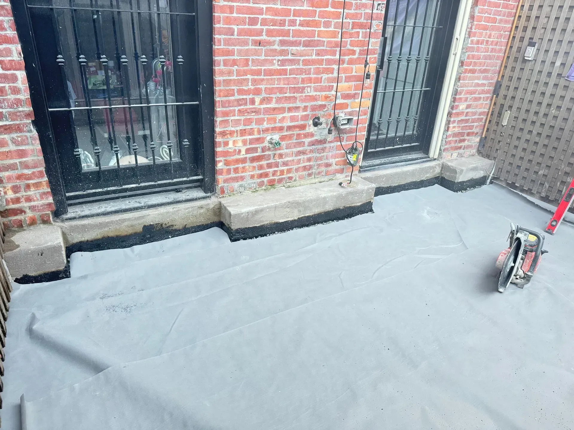 A gray waterproof membrane covers a brick building's terrace. Two barred doors and a tool case are visible.