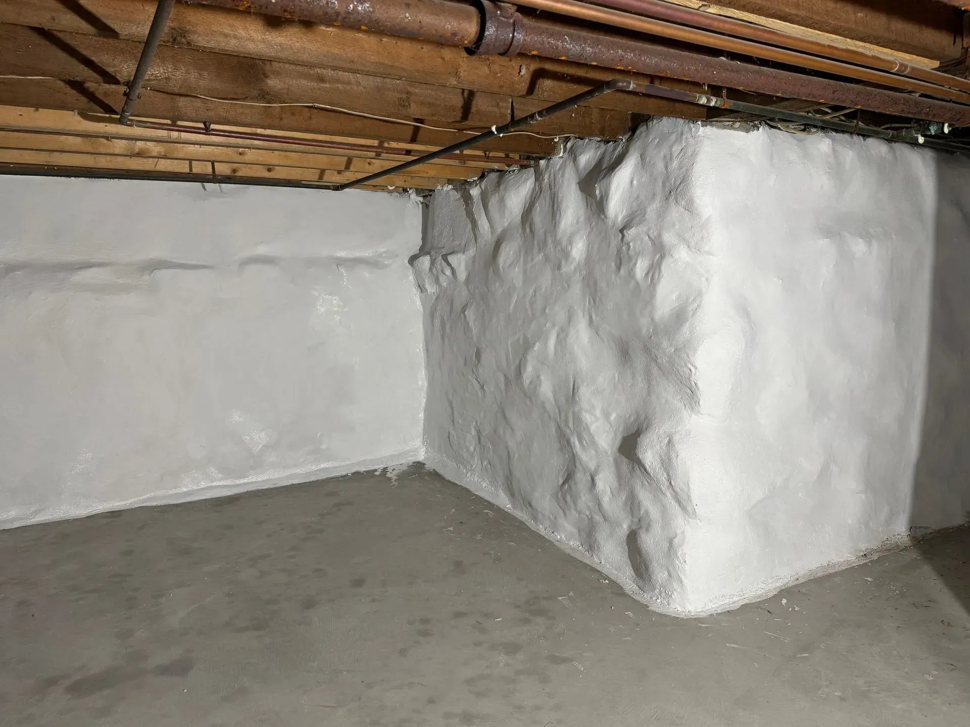 Basement corner with white-painted, textured concrete walls, wooden ceiling beams, and a concrete floor.