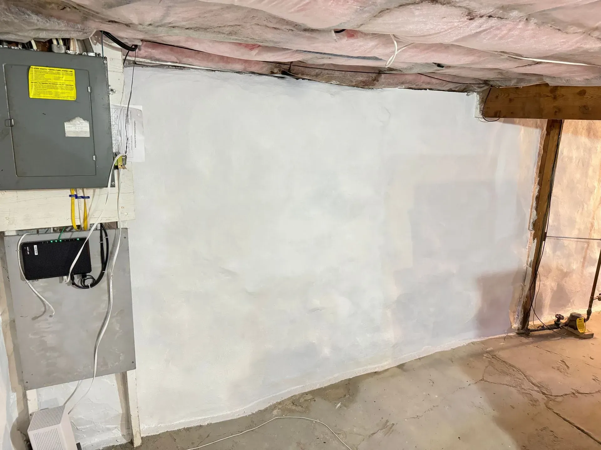 Basement wall with electrical panel and insulated ceiling. White wall, gray panel, exposed wood beam.