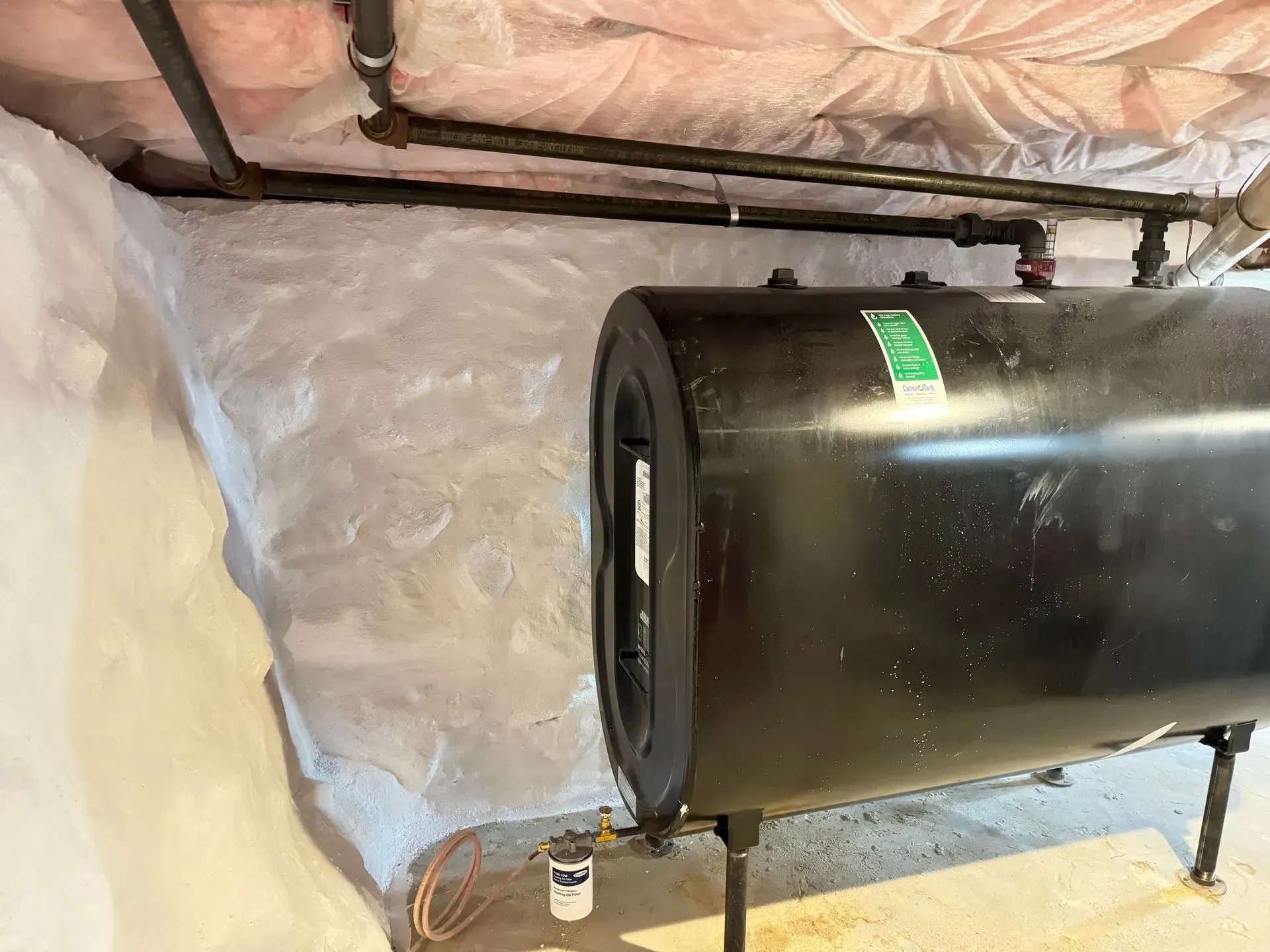 Black oil tank in a basement, next to a textured white wall. Pipes and insulation are visible overhead.