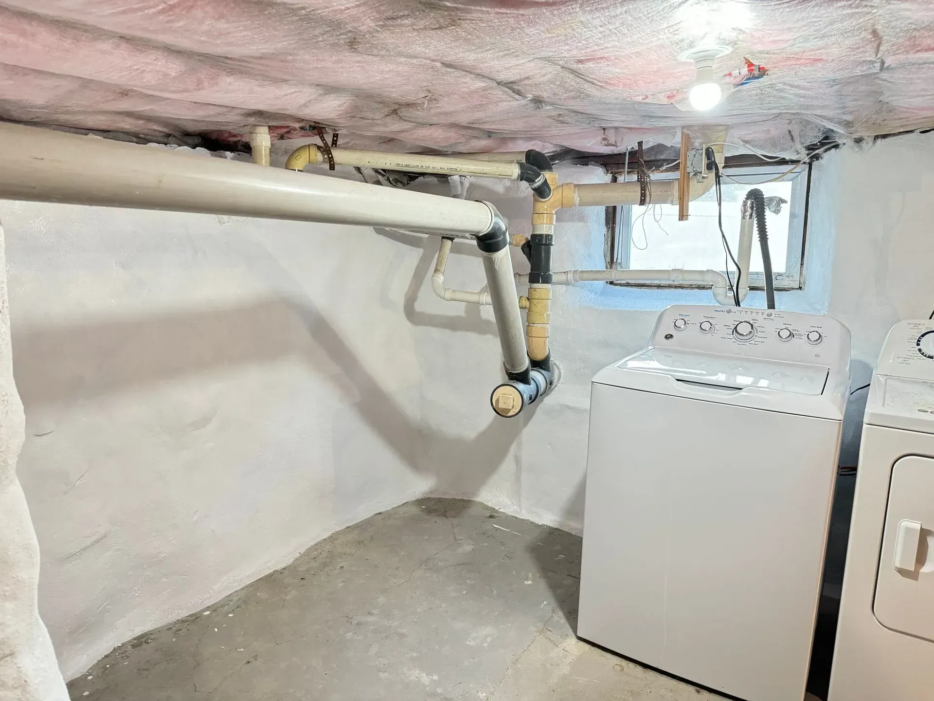 Basement laundry area with washer and dryer, pipes, and exposed ceiling insulation. White appliances against white walls.