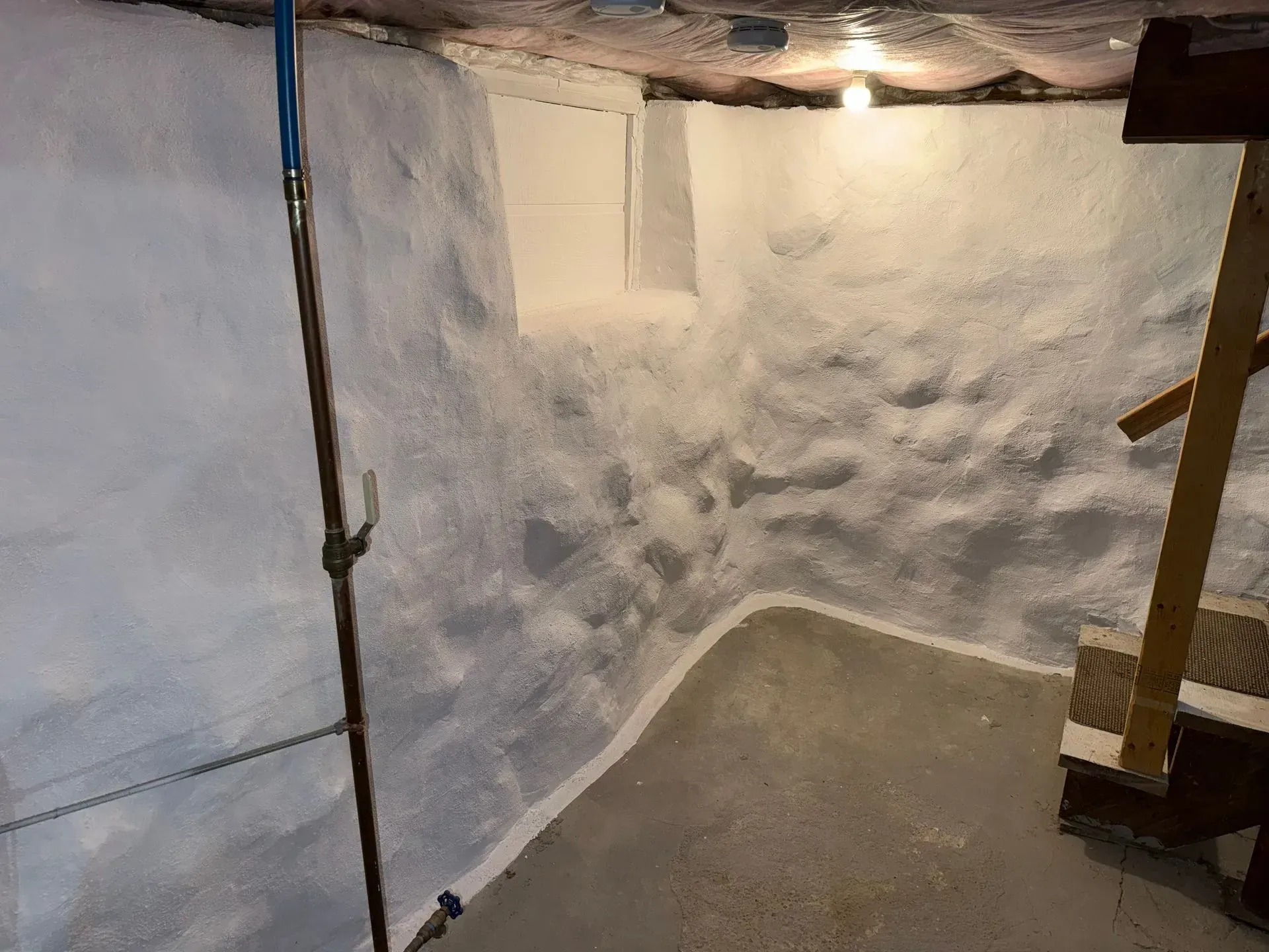 Basement corner with painted stone walls, concrete floor, and wooden stairs. Light bulb visible.