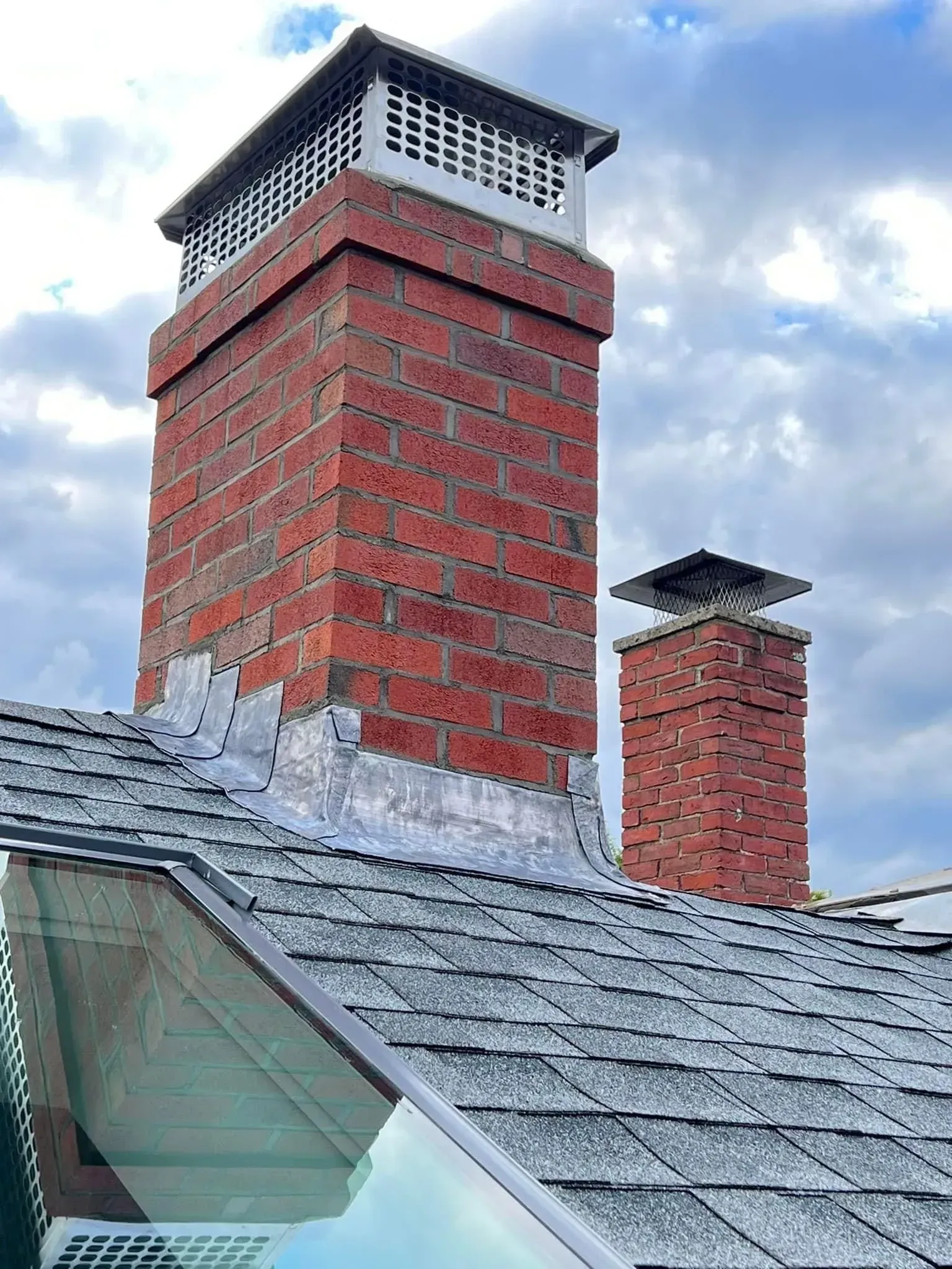 Two brick chimneys on a gray shingled roof, topped with metal caps, against a cloudy sky.