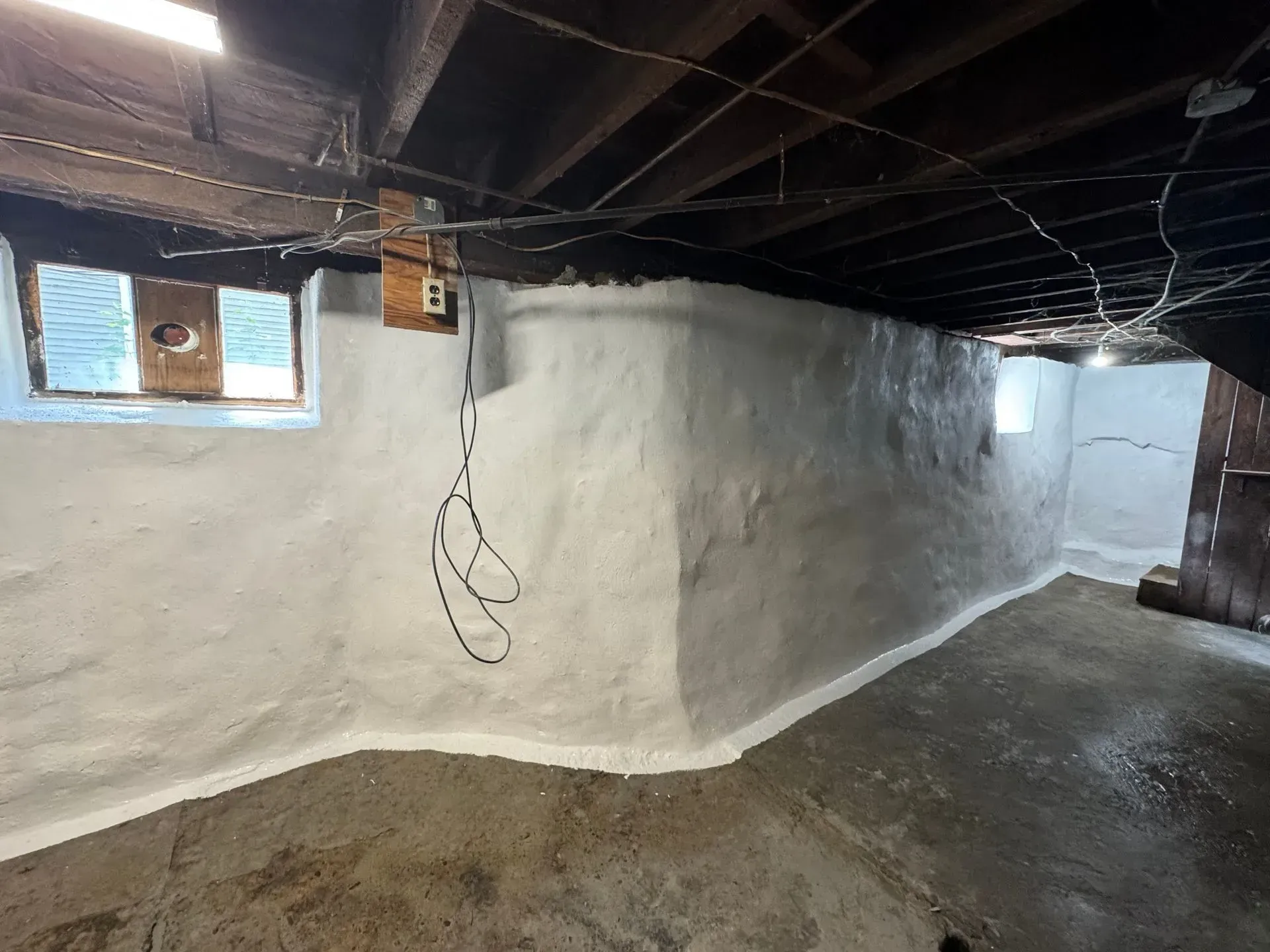 Basement interior, white-painted stone walls, cement floor, wooden beams ceiling, small windows, electrical wiring.