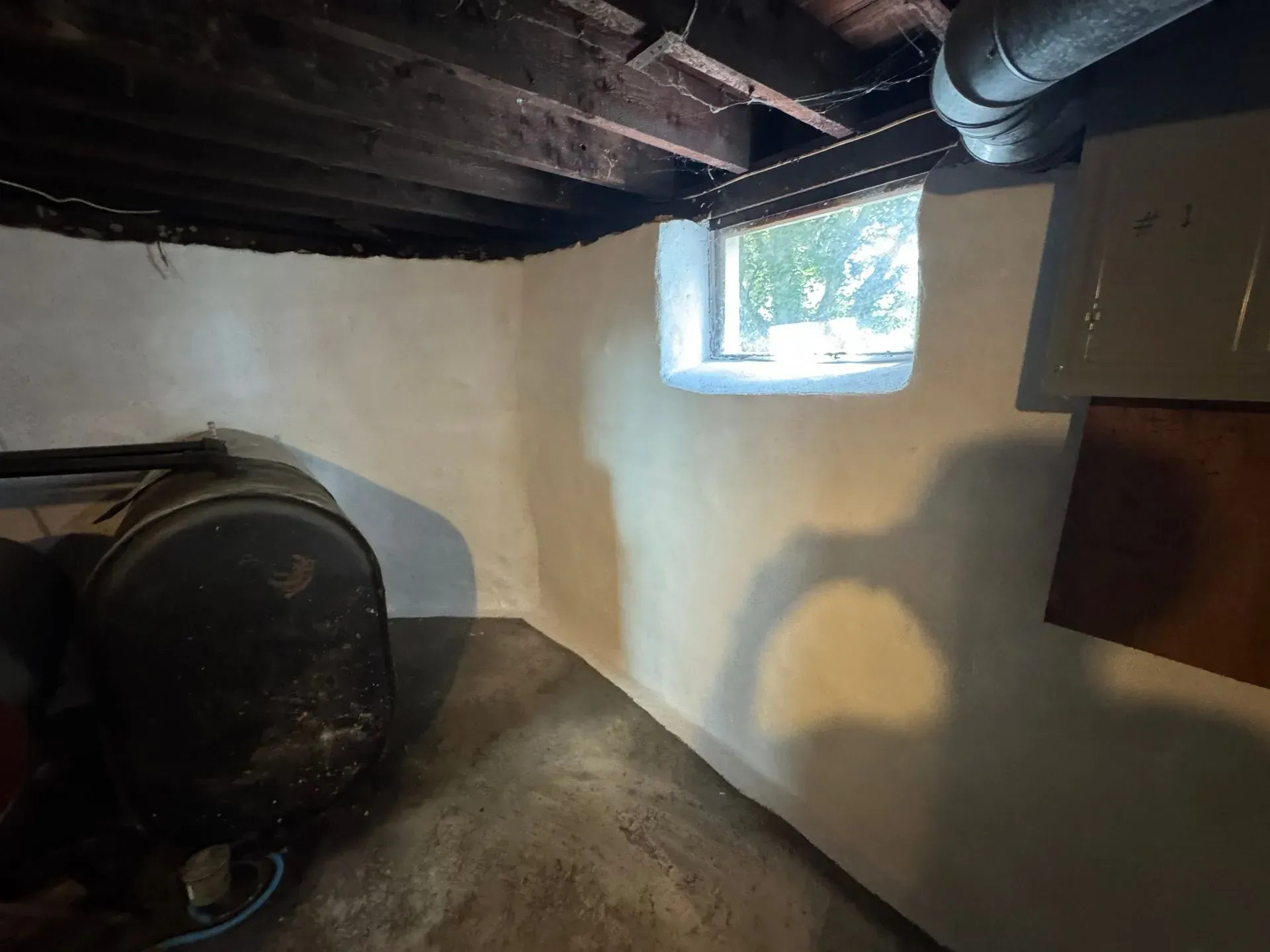 Basement corner with white-painted walls, small window, dark tank, exposed beams, and concrete floor.