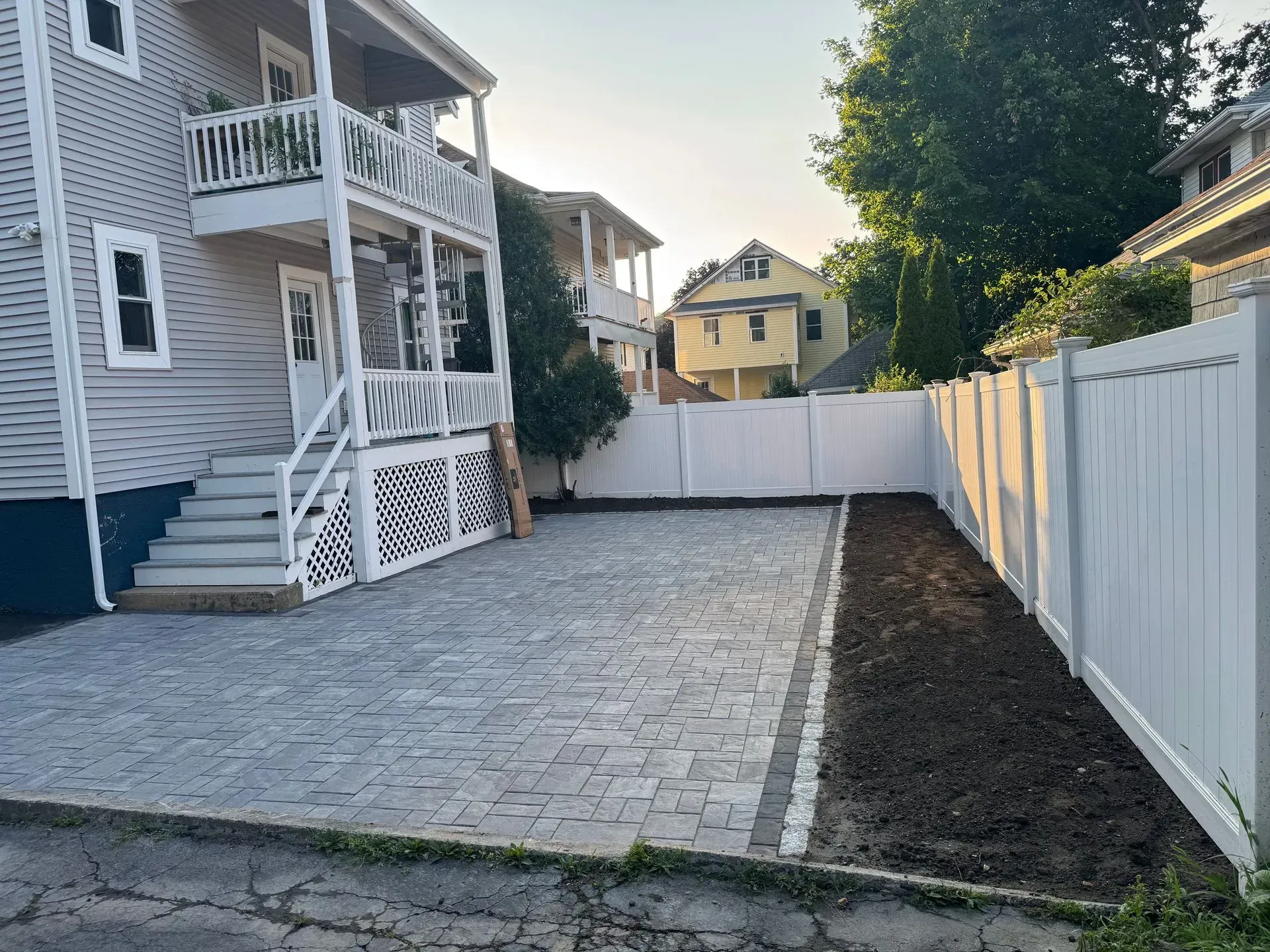 Backyard paved with gray pavers, white fence, and building. Garden bed with dark soil along the fence.