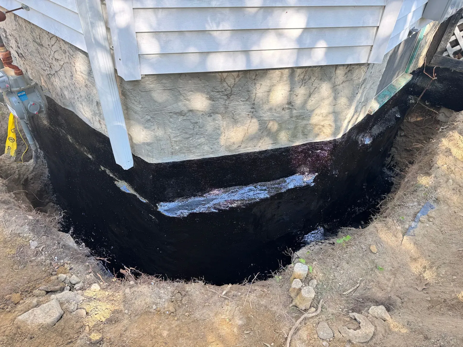 Dark, asphalt-coated trench around the base of a light-colored house. Earth and debris surround.