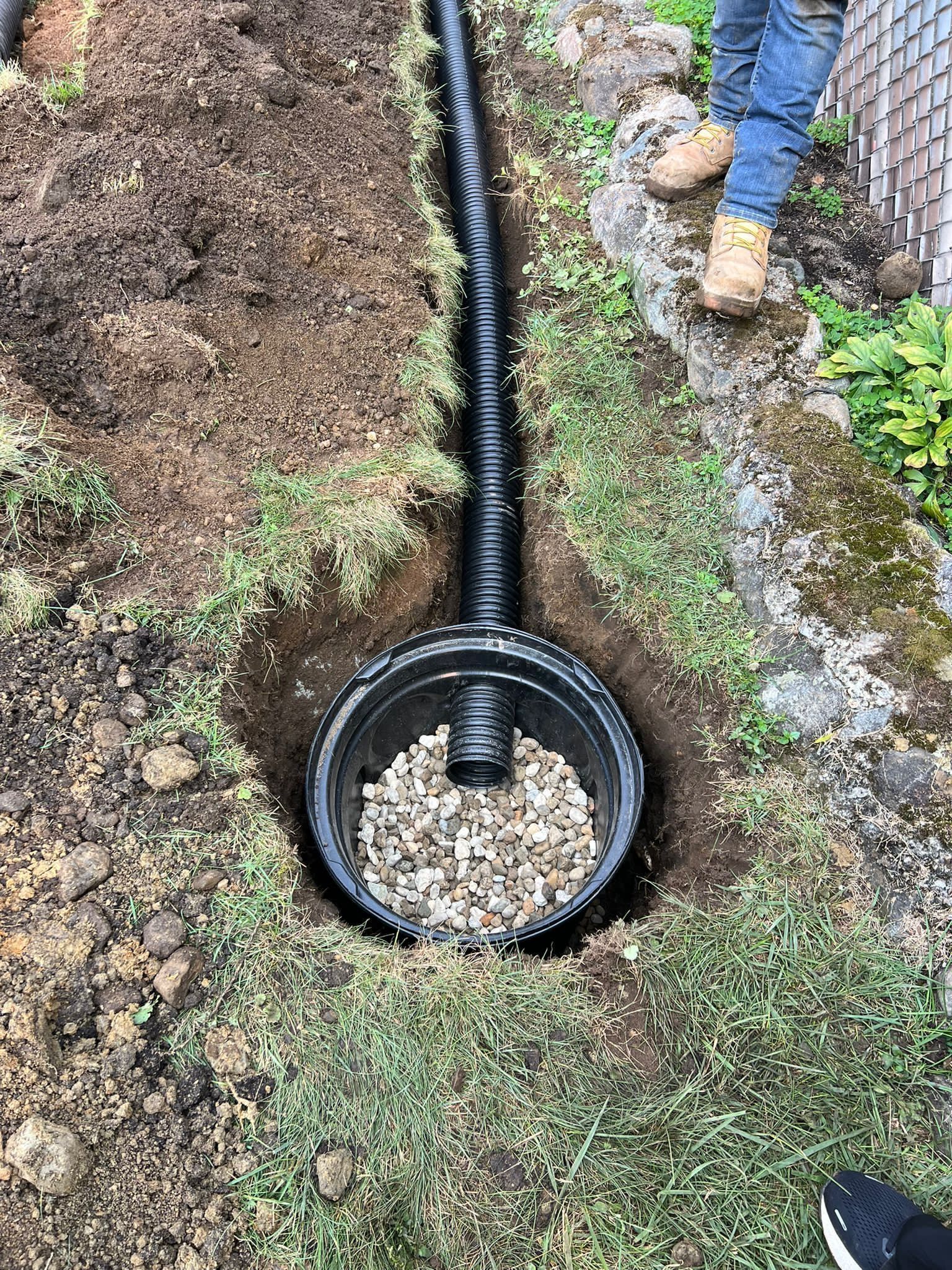 Black drainage pipe and basin with gravel in a trench, near a person's legs.