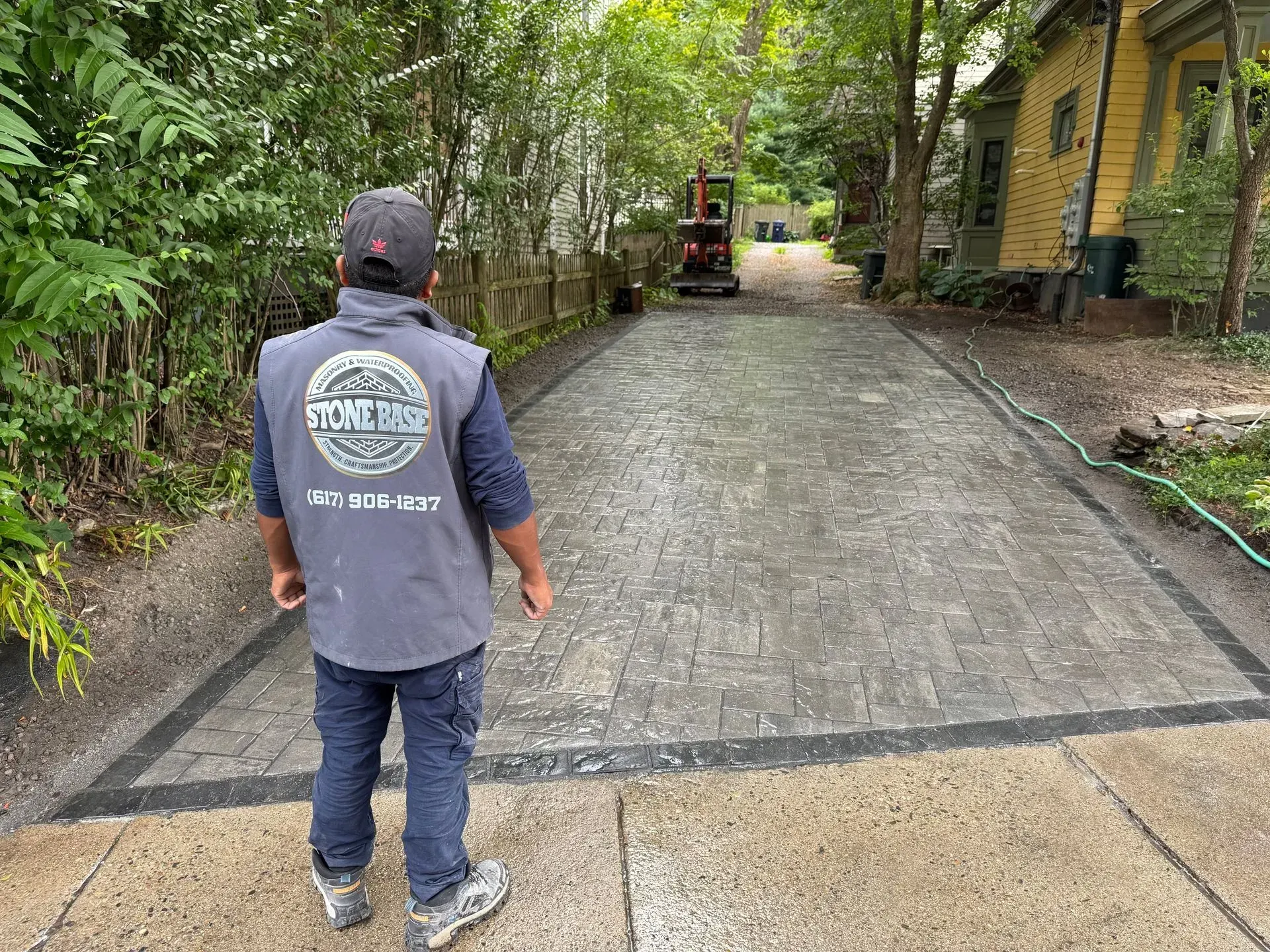 Man in work vest surveys a brick paver driveway under construction, with a small excavator in the distance.