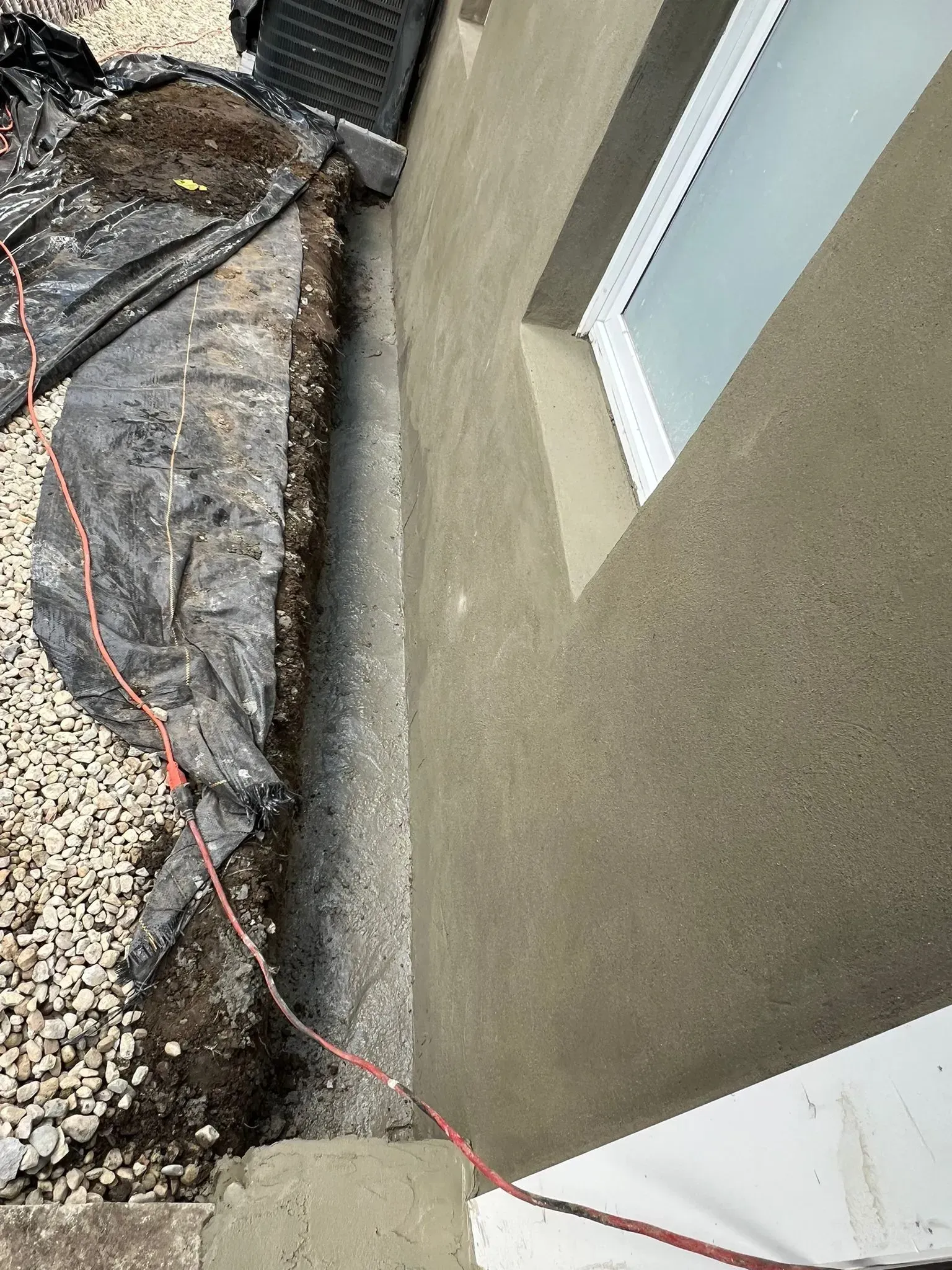 A narrow trench next to a building wall with a window, covered in black sheeting and gravel.