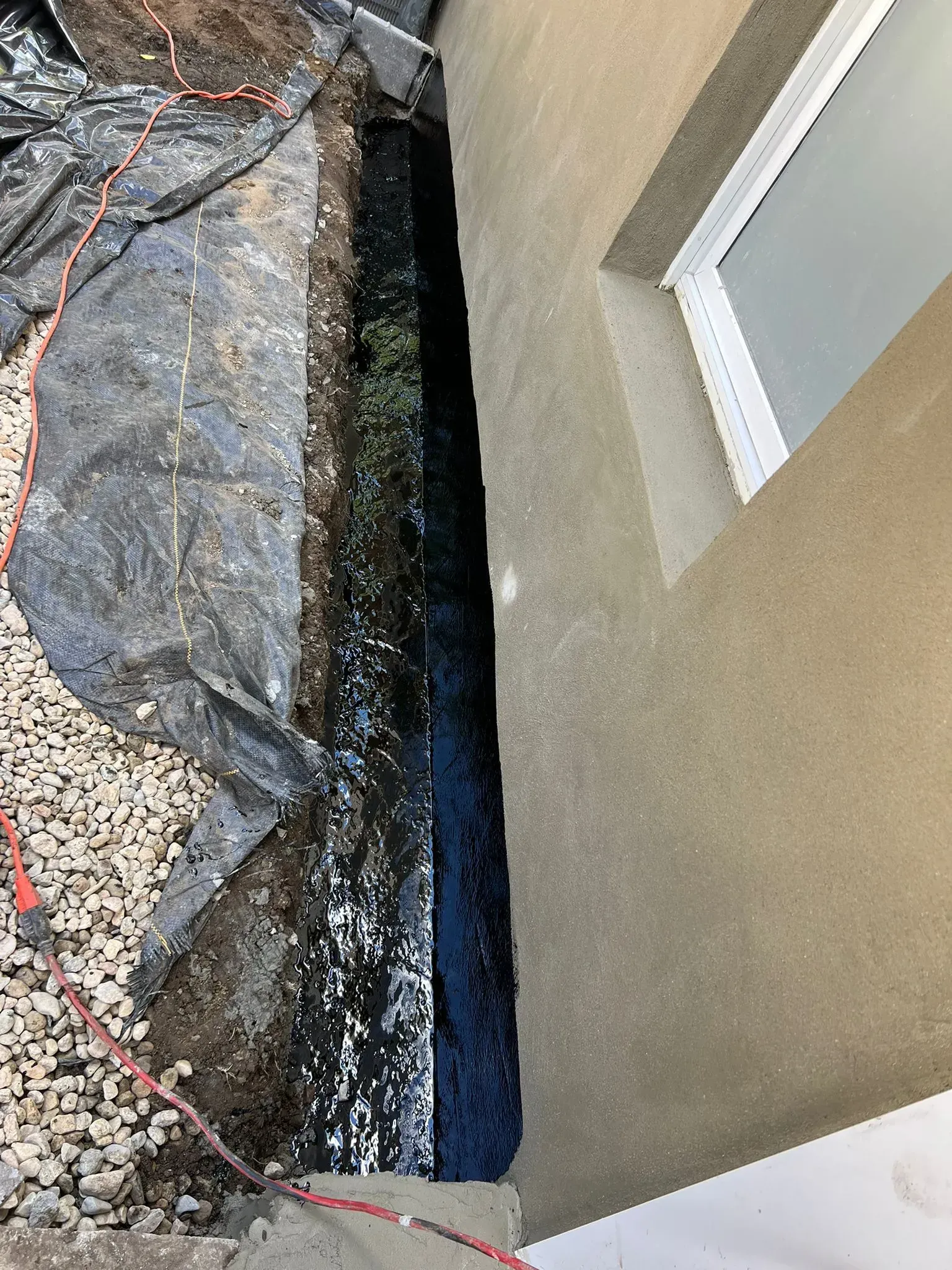 A narrow trench with black sealant alongside a building's stucco wall and window. Gravel and plastic sheeting are also present.