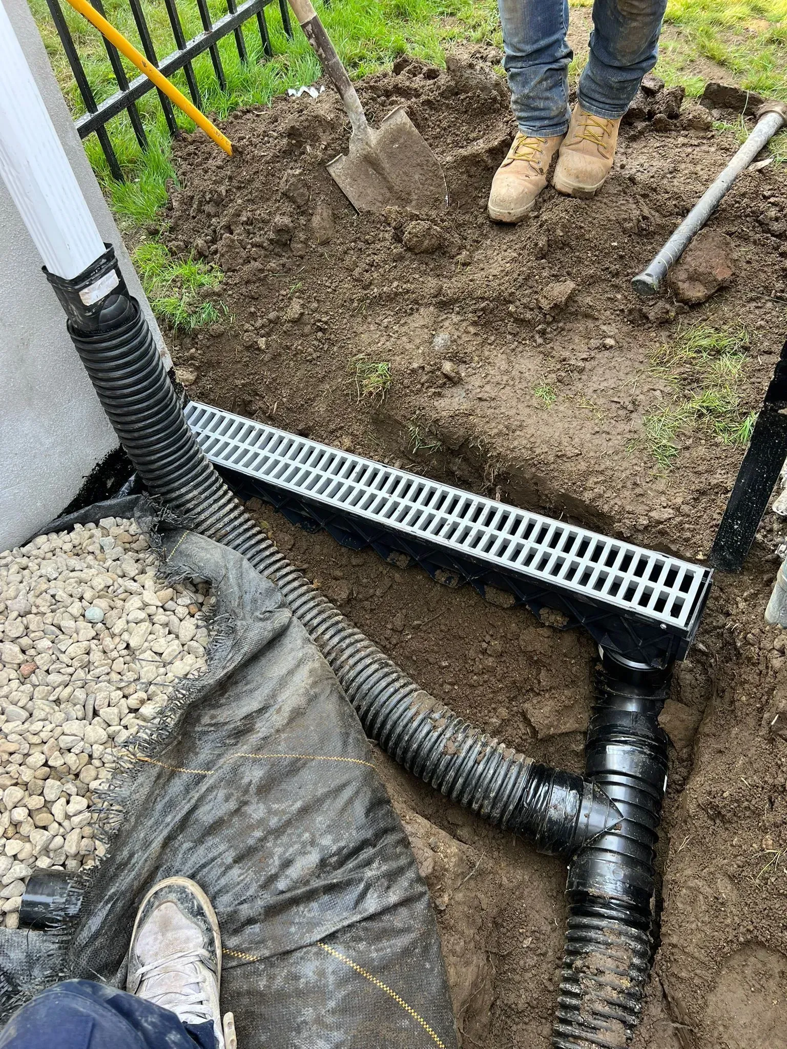 Drainage system installation in progress: black pipe and grate in a trench. Person in boots stands nearby.