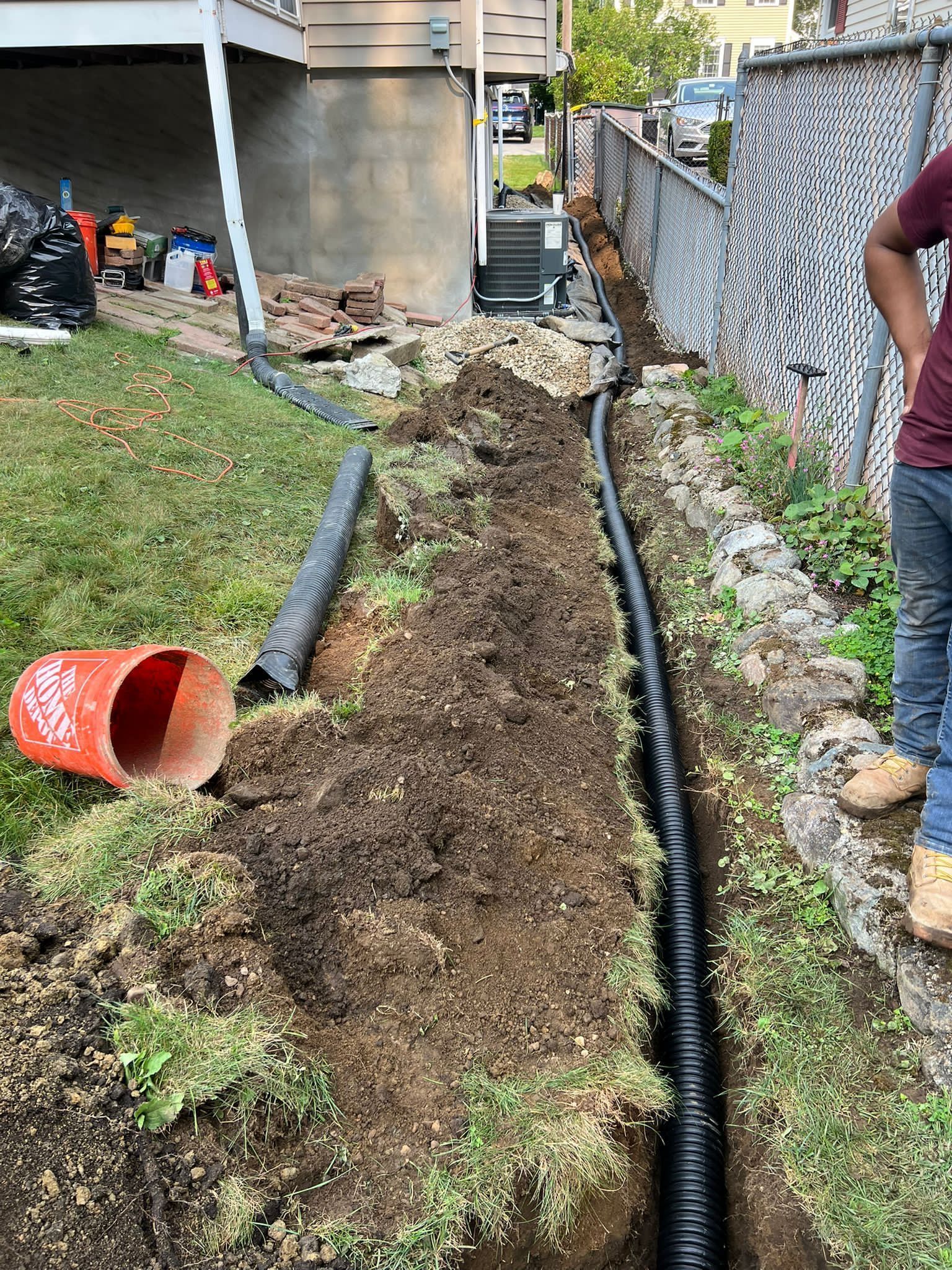 Trench with black drainage pipe being installed alongside a house, fence, and a person.