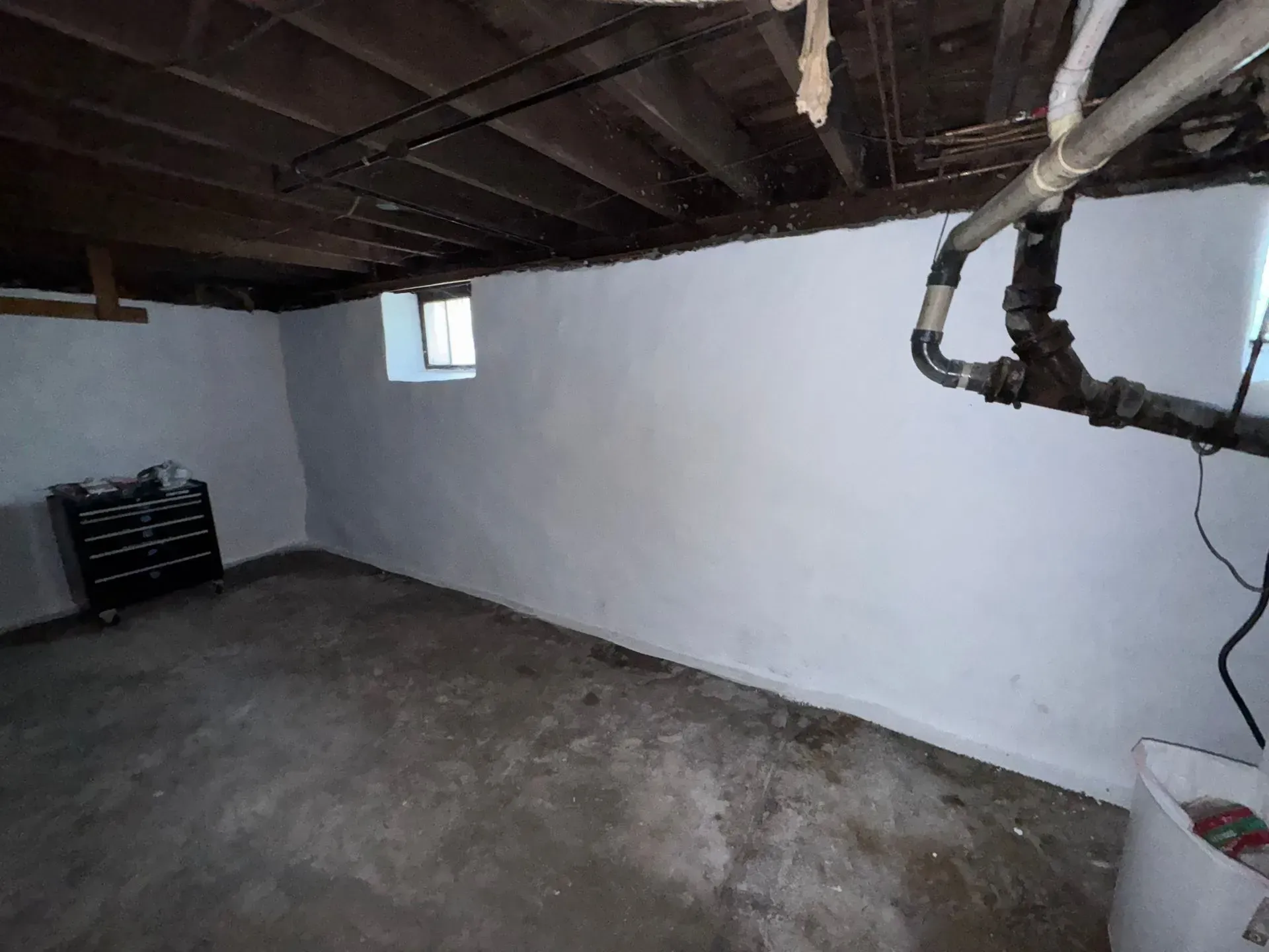 Basement with painted white walls, concrete floor, pipes, small window, and tool chest.