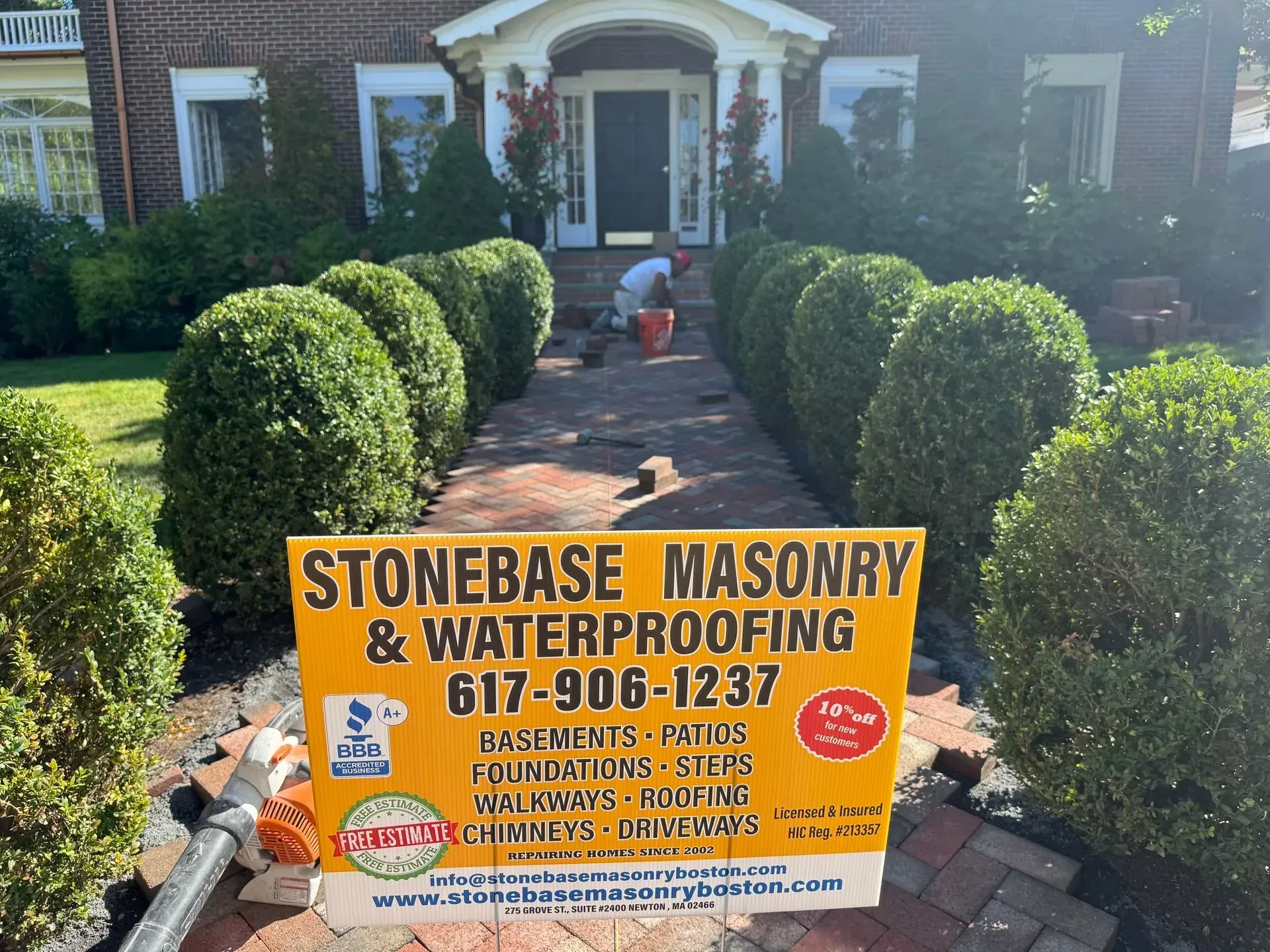A person works on a brick walkway in front of a brick house. A sign for Stonebase Masonry is in the foreground.