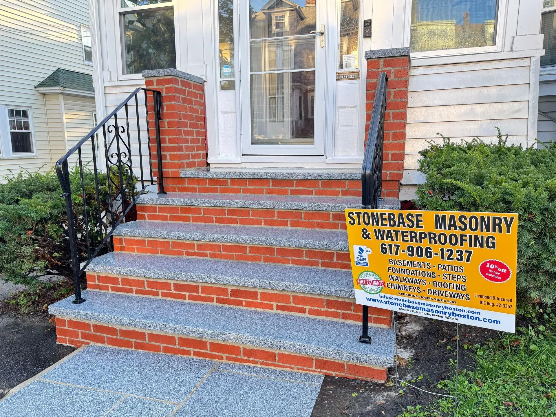 Brick and stone steps leading to a house entrance, with black handrails and a sign for a masonry company.