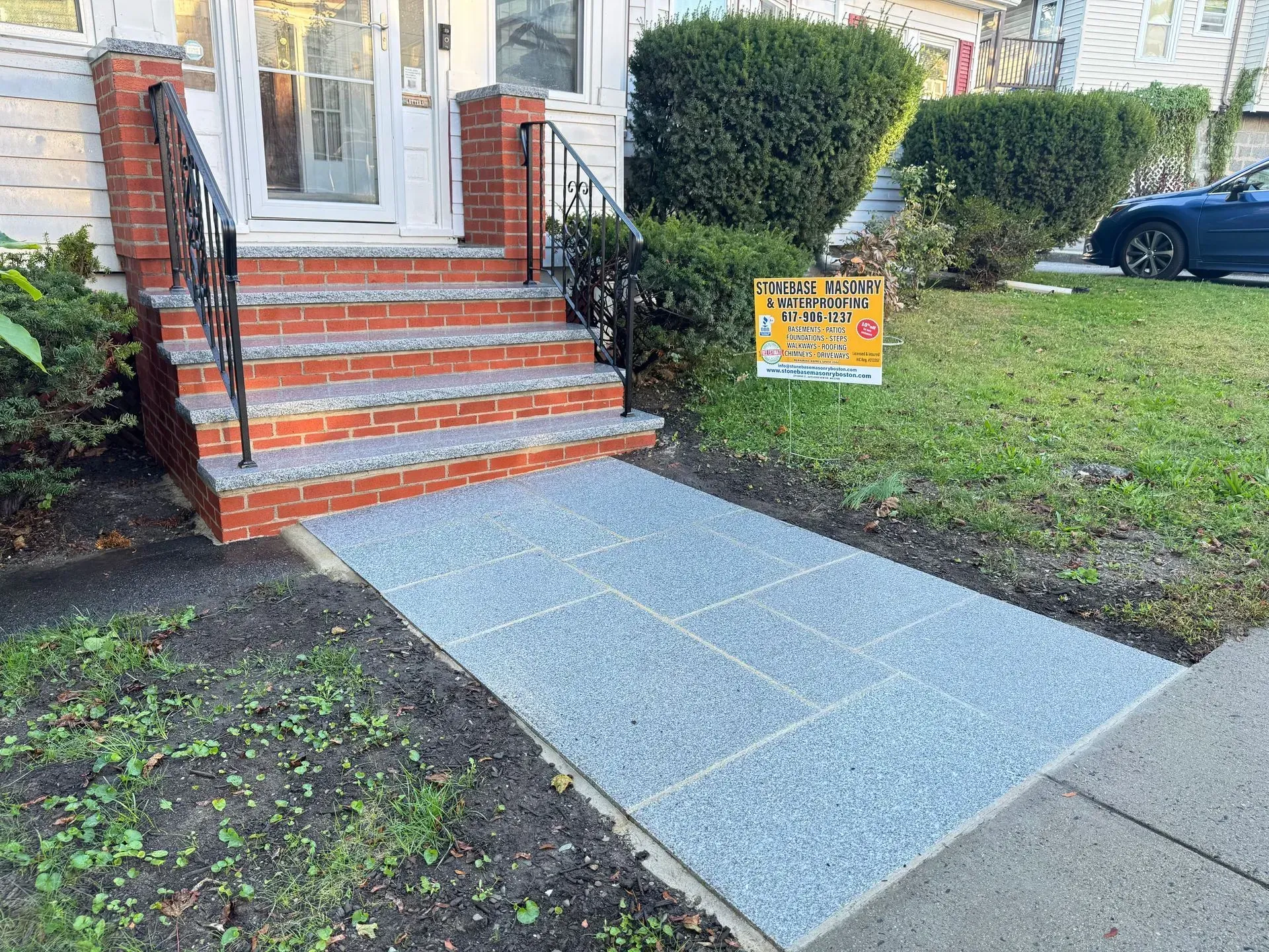 Grey stone walkway and brick steps leading to a house entrance. Green lawn, bushes, and a parked car are in the background.