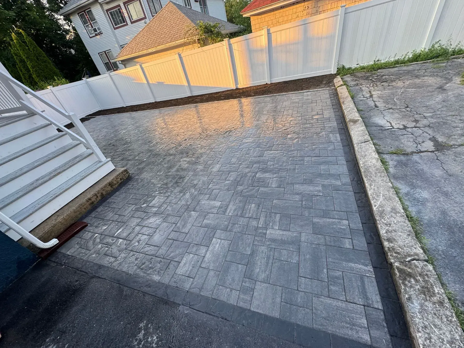Paver driveway next to stairs and a white fence, bathed in sunlight.