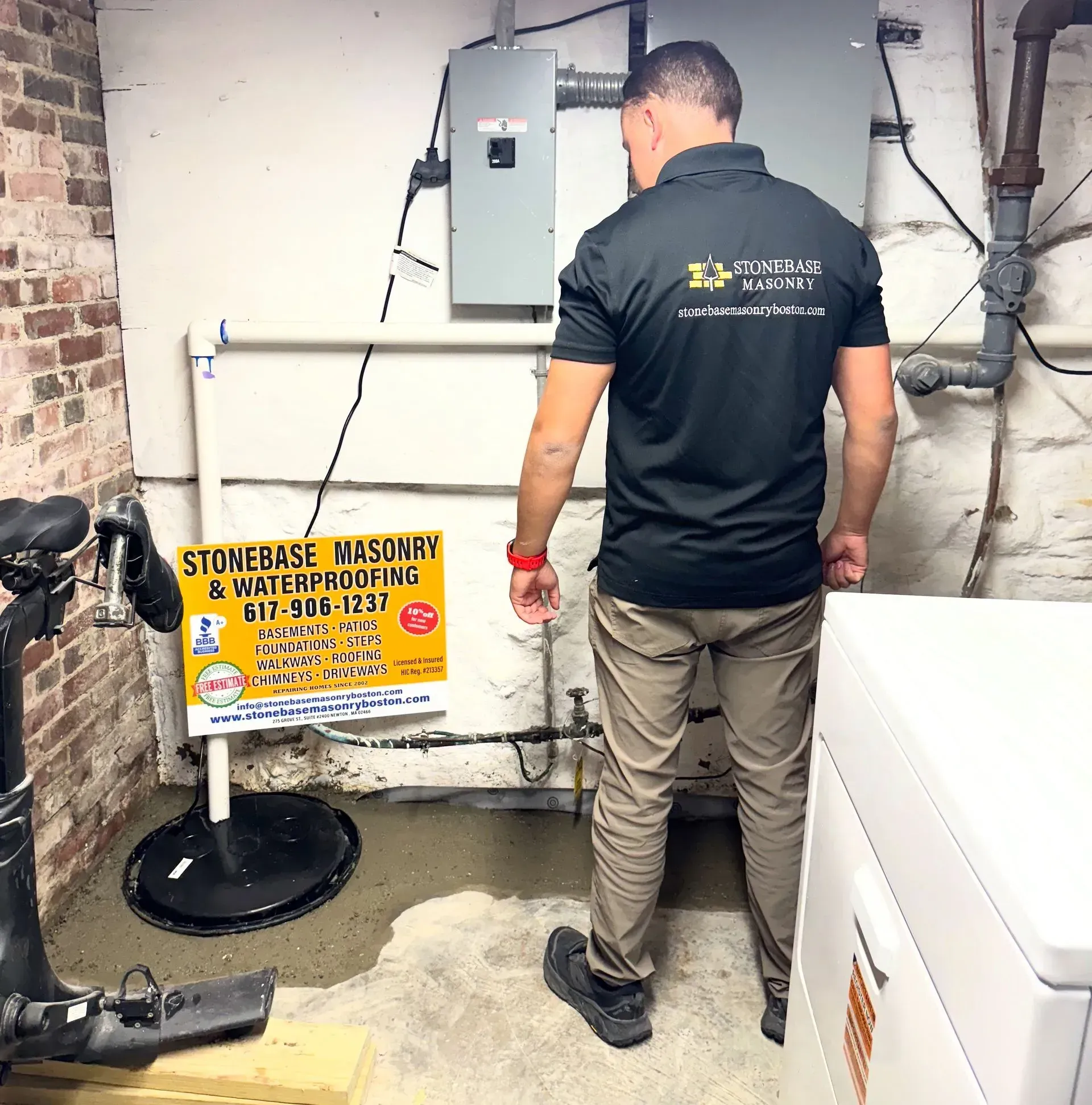 A person in a black polo shirt examines a basement space with a Stonebase Masonry sign, concrete floor, and pipes.