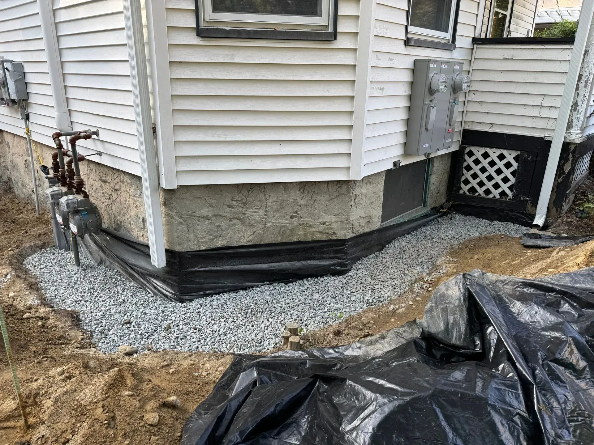 Gravel drainage around the foundation of a white house. Black plastic liner and electrical box are visible.