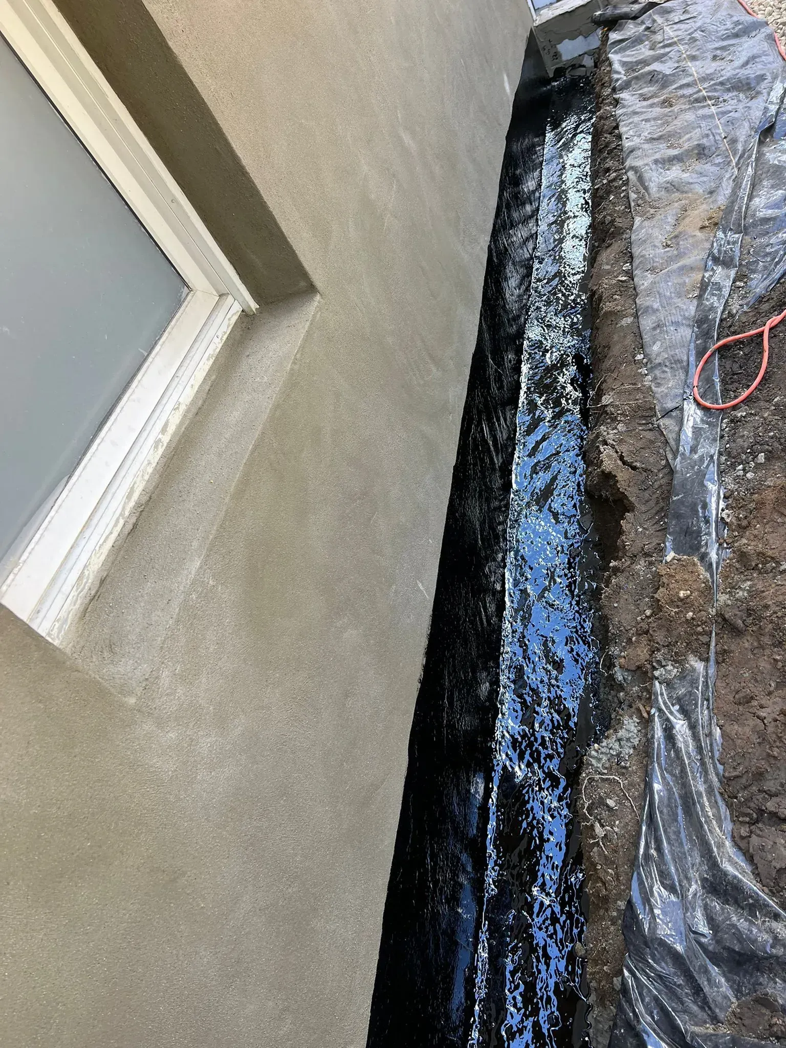 Exterior wall next to a trench filled with black material, potentially waterproofing. Dark plastic sheeting on the ground.