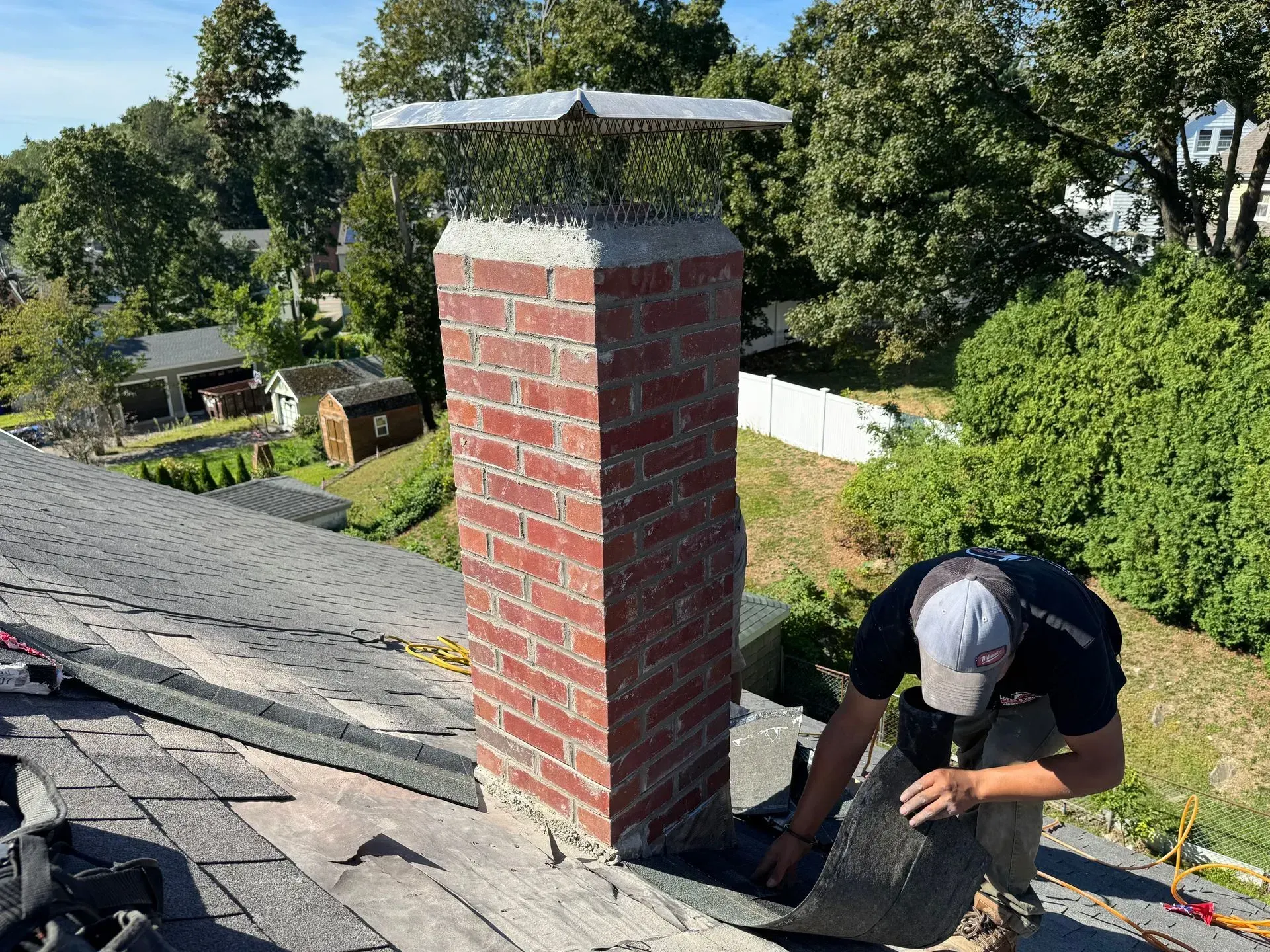 Roofer working on flashing around a brick chimney on a shingled roof, trees in the background.