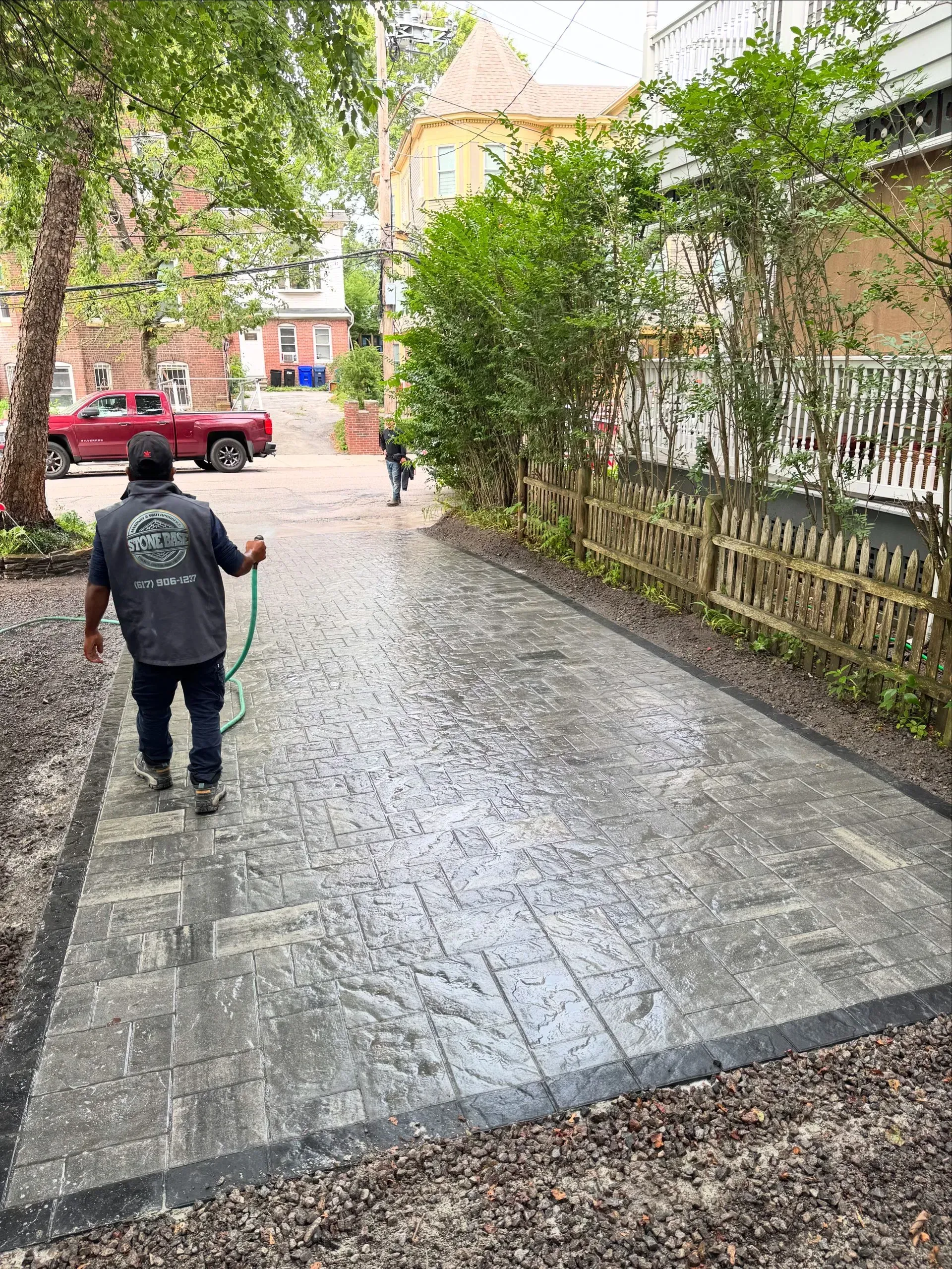 Man spraying a wet, patterned concrete driveway with a hose, in a narrow space between buildings.