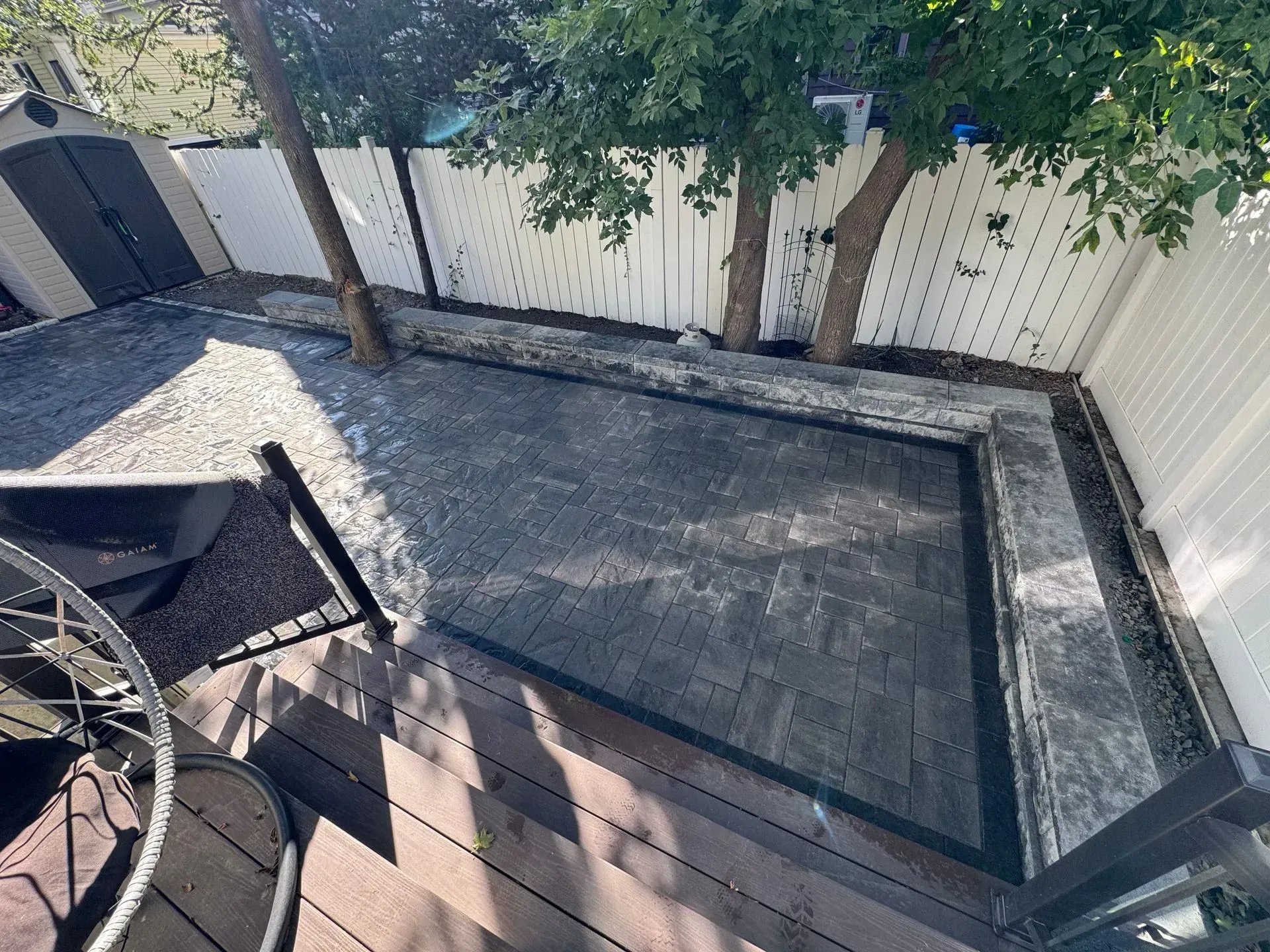 Backyard patio with a dark brick area, trees, white fence, and shed.