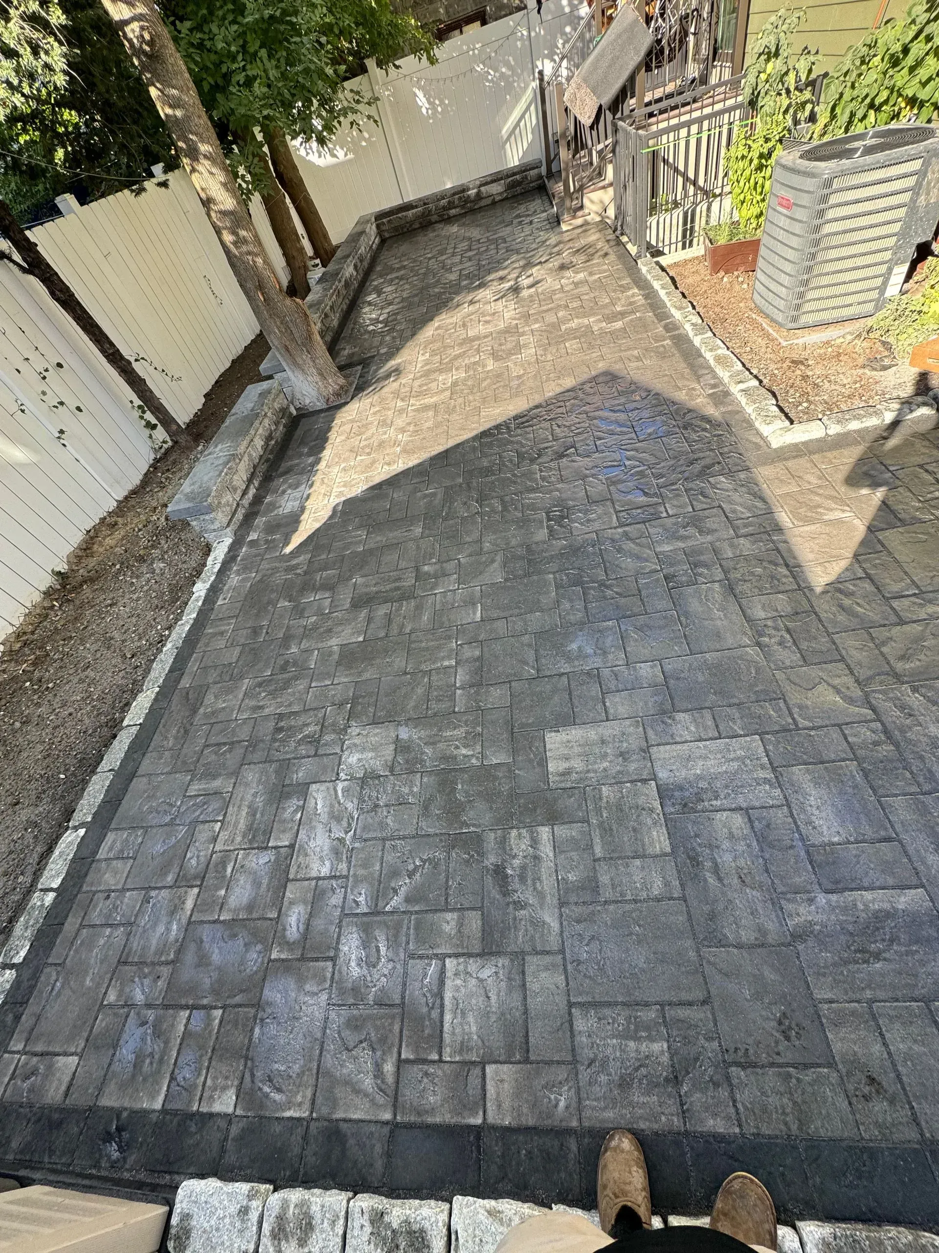 Dark gray paved patio with a tree, white fence, and person's feet.