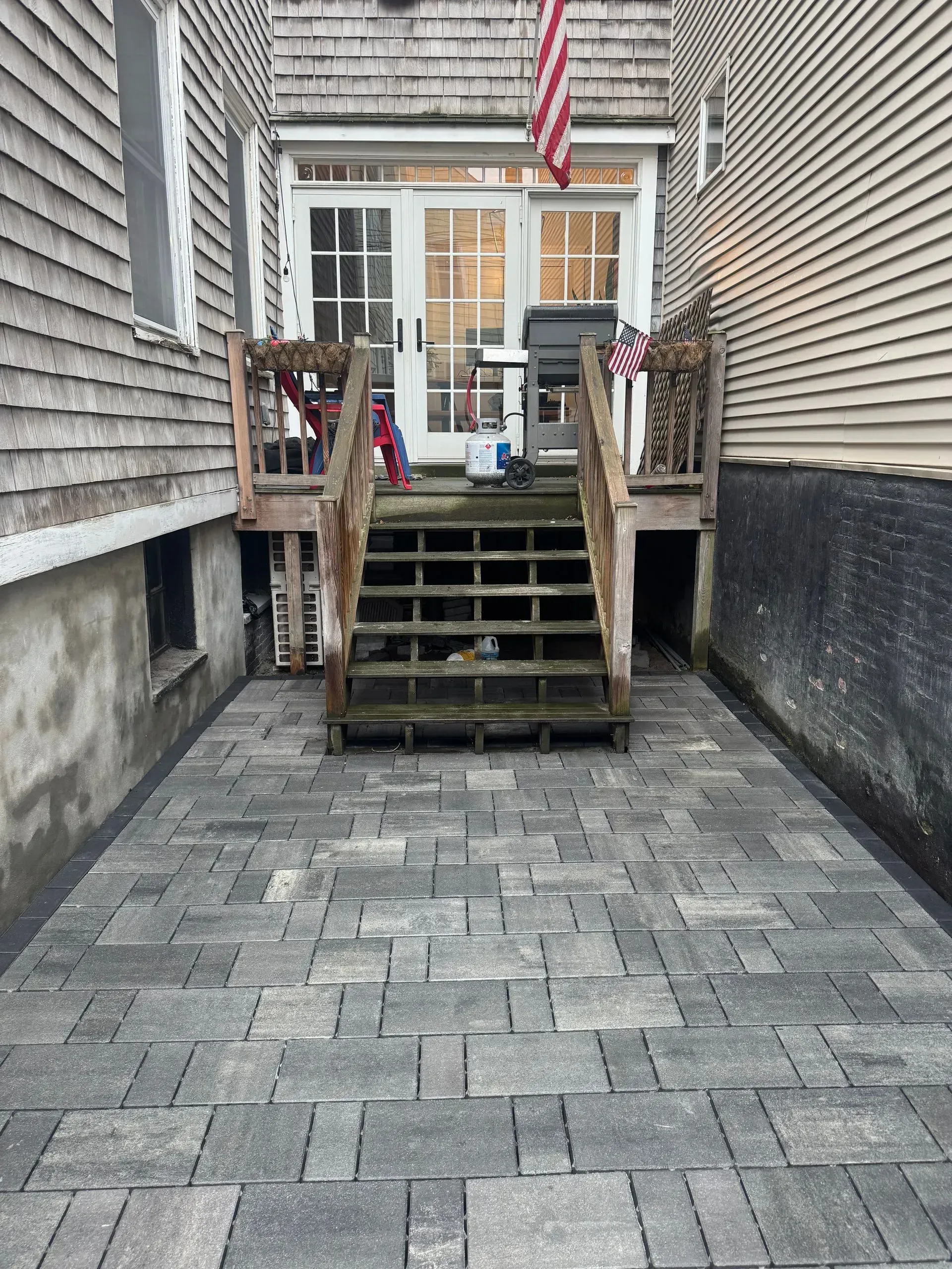 Stone patio leads to wooden stairs and a doorway with an American flag. Buildings flank the narrow space.