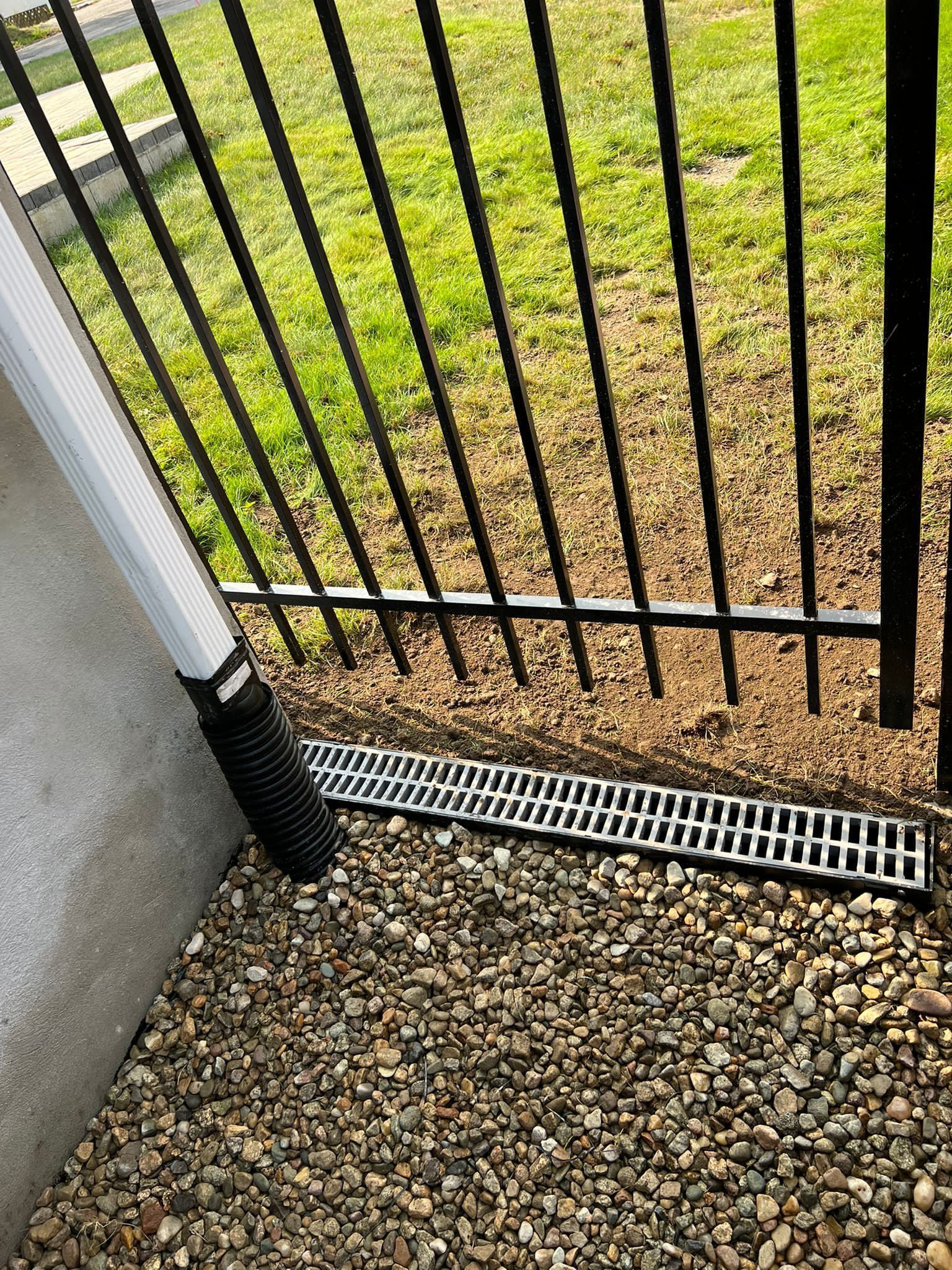 Black metal fence next to a gravel area with a linear drain and a white downspout.