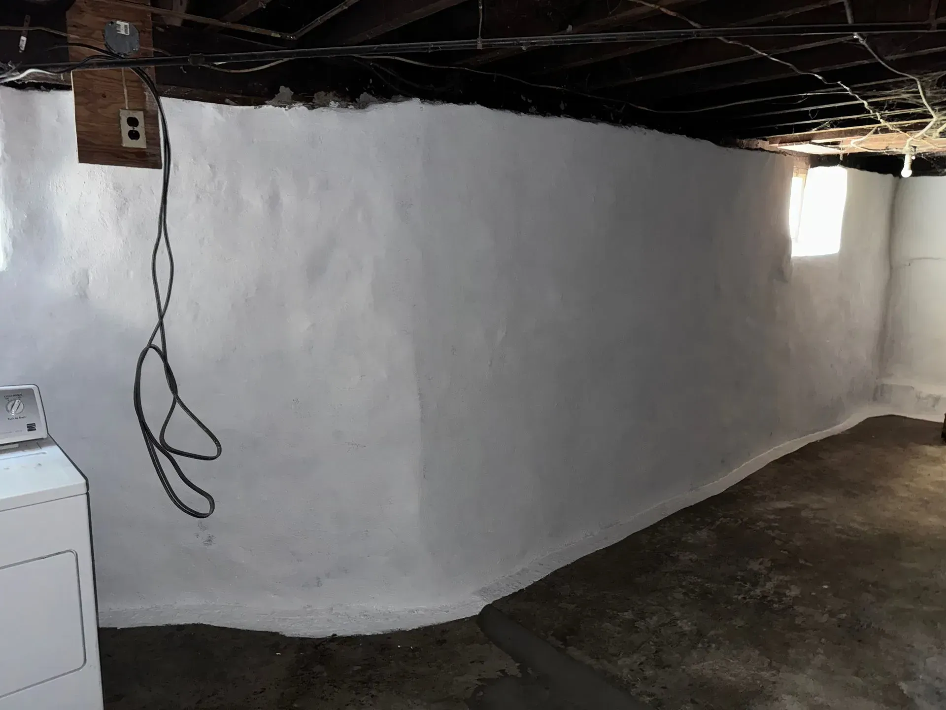 White painted basement wall with window and laundry machine in a concrete room.