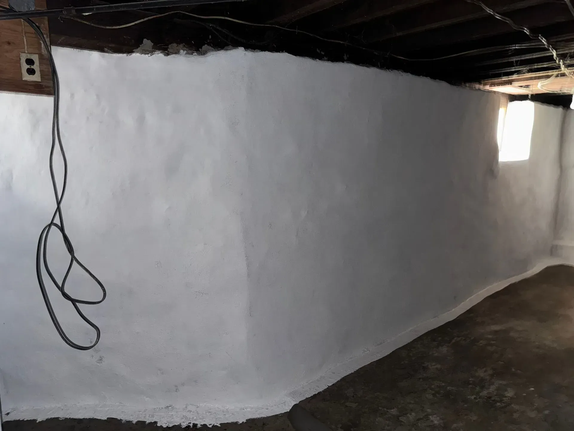 White-painted basement wall with a looping black cable hanging on the left and a small window on the right.