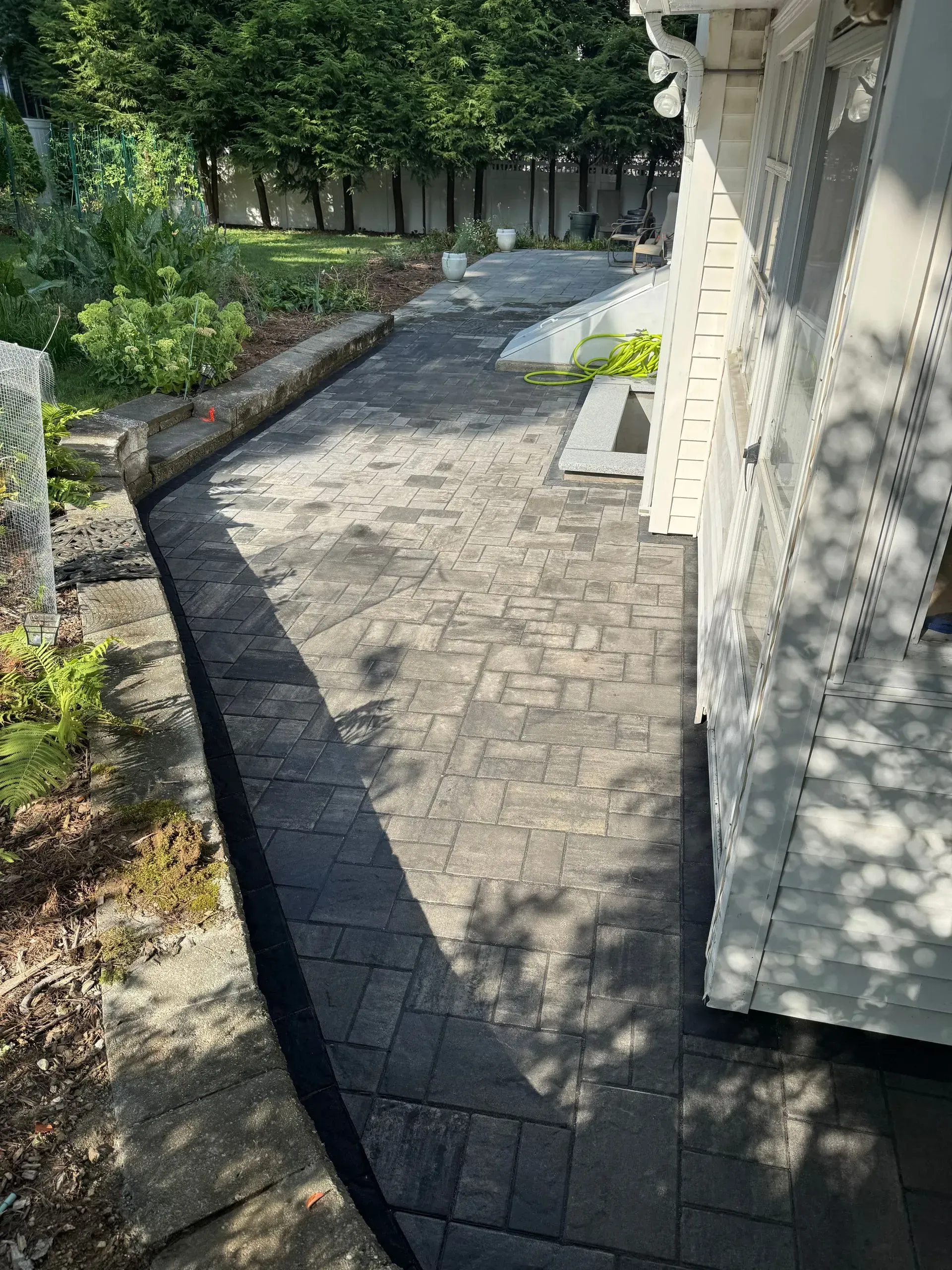 Brick patio next to a white house, leading to a garden area, with trees in background.