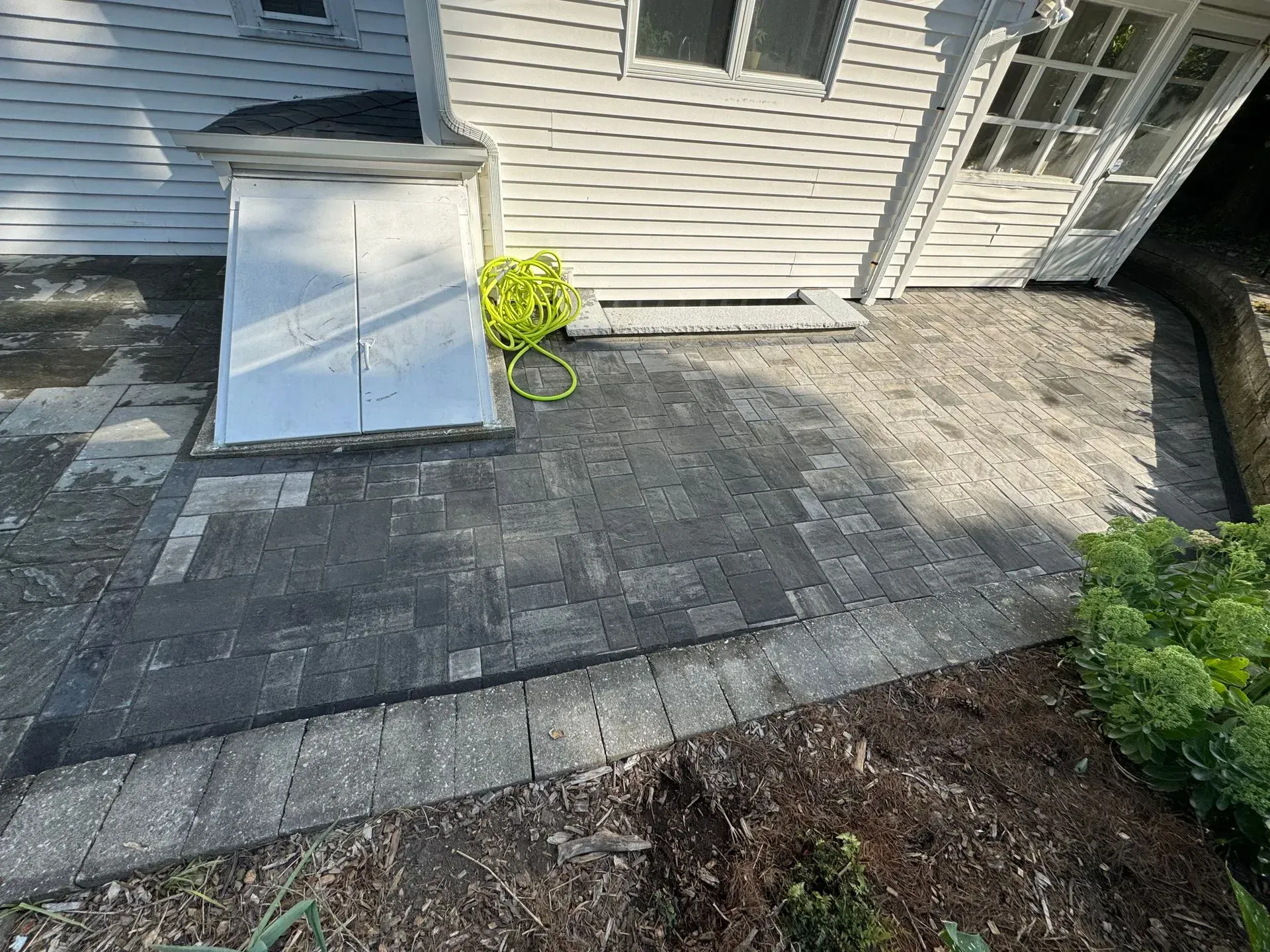 Brick patio next to a white house with a basement door and a garden hose.