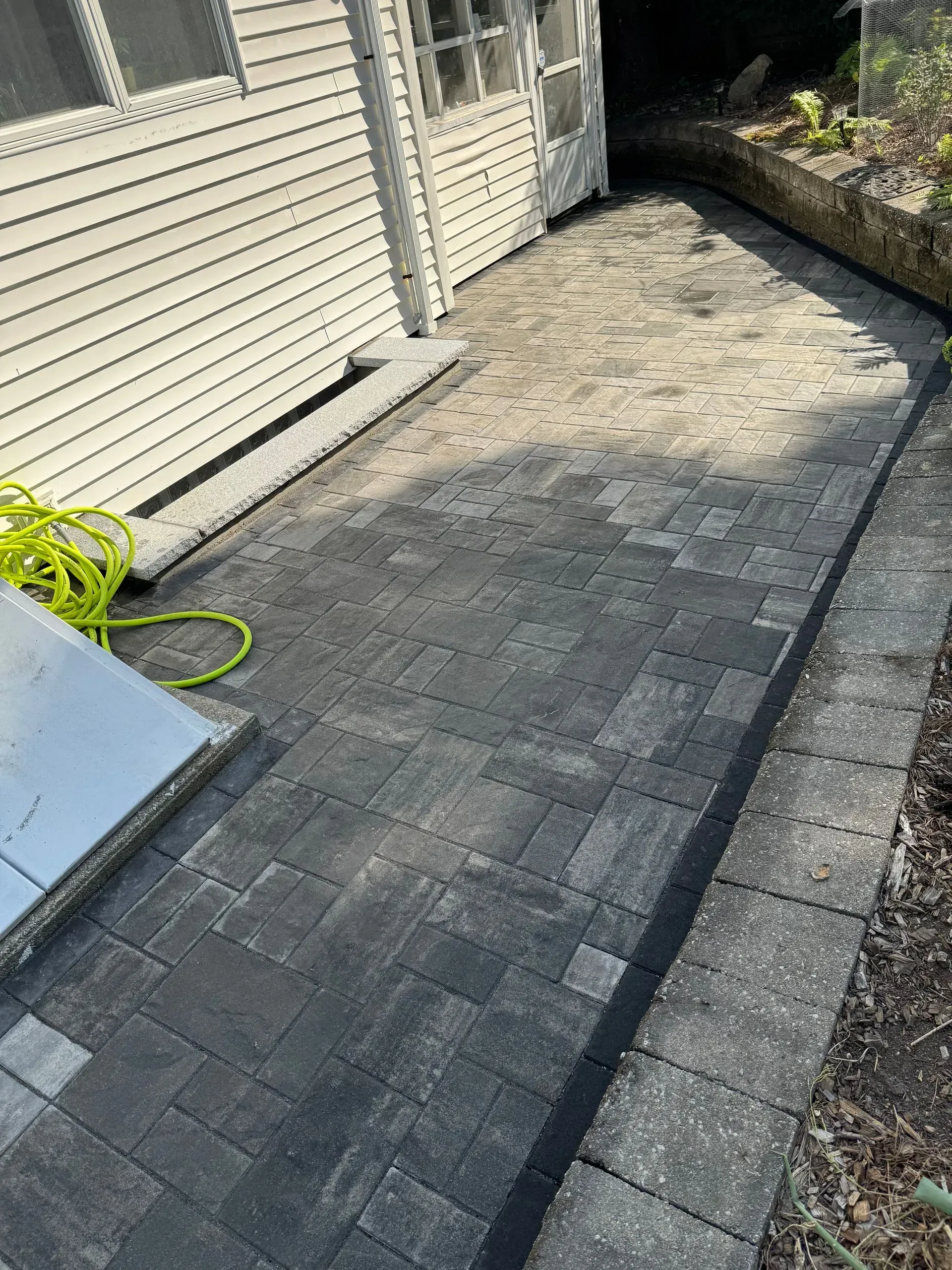 Brick patio with dark gray pavers next to a house with beige siding and a stone retaining wall.