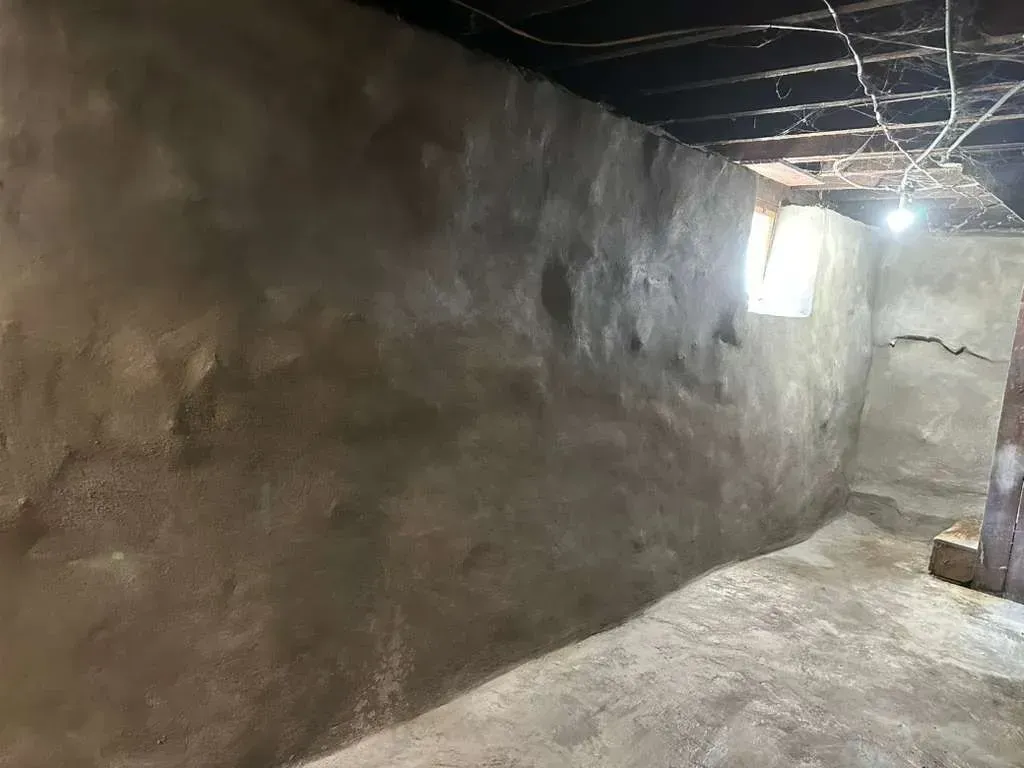 Gray concrete basement with rough walls, small window, and exposed wooden ceiling beams.