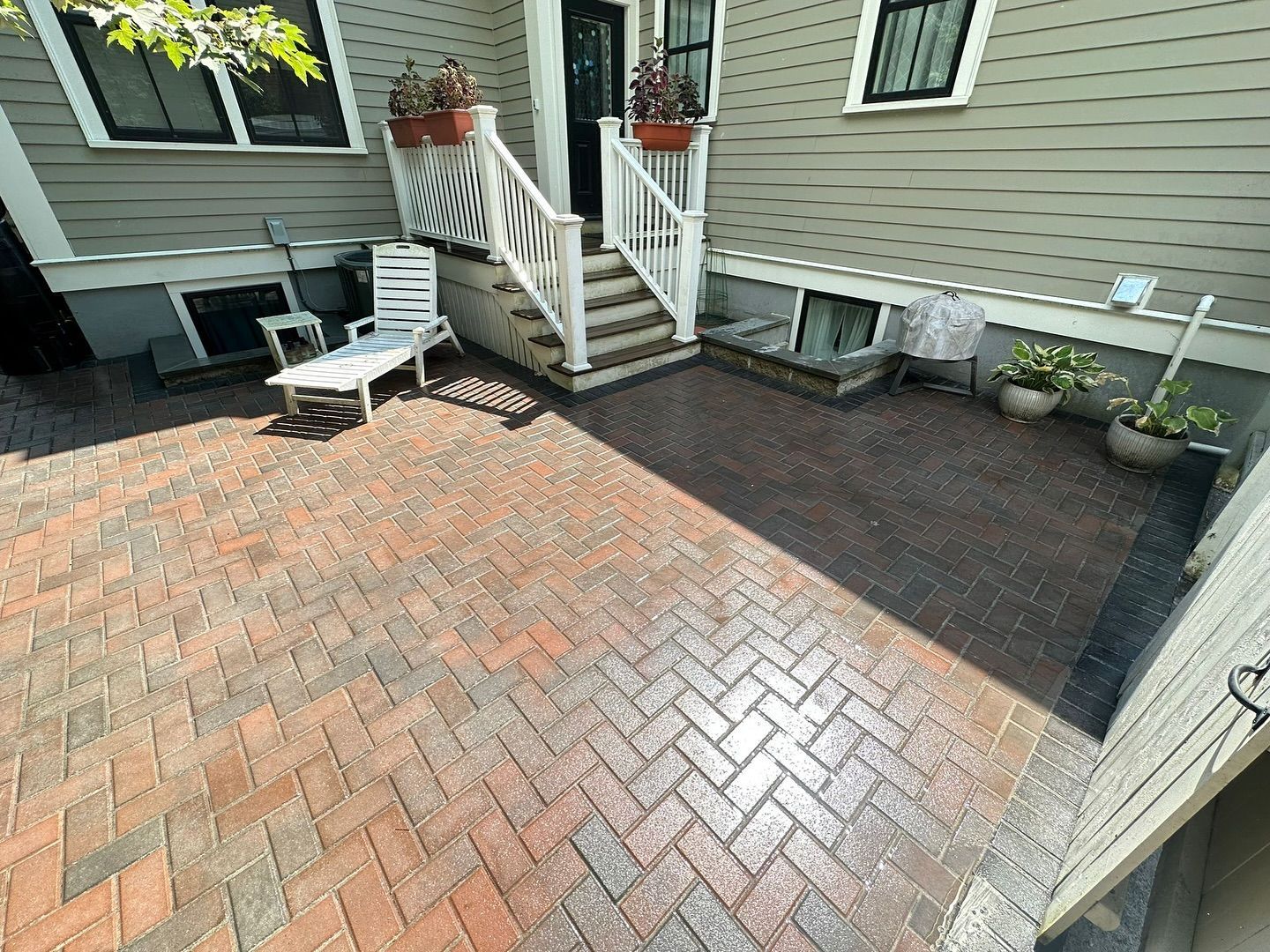 Brick-paved patio beside a house with white stairs, outdoor chairs, and planter boxes in sunlight