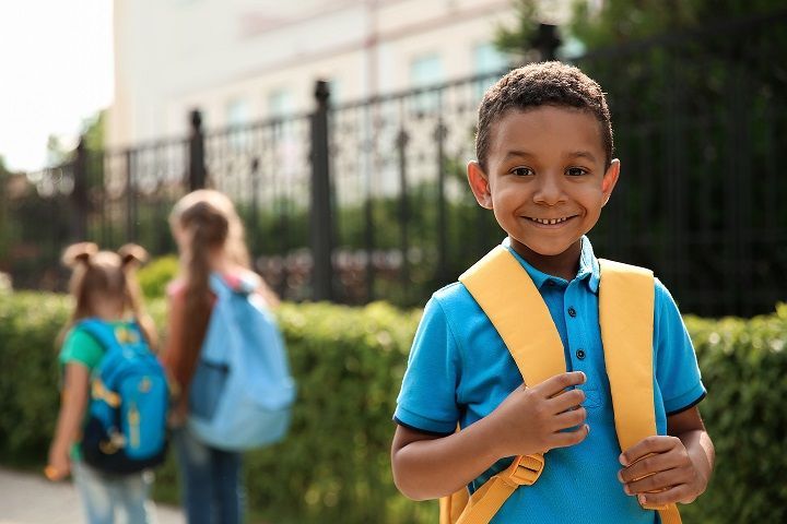 A young boy with a yellow backpack is standing in front of a school building.